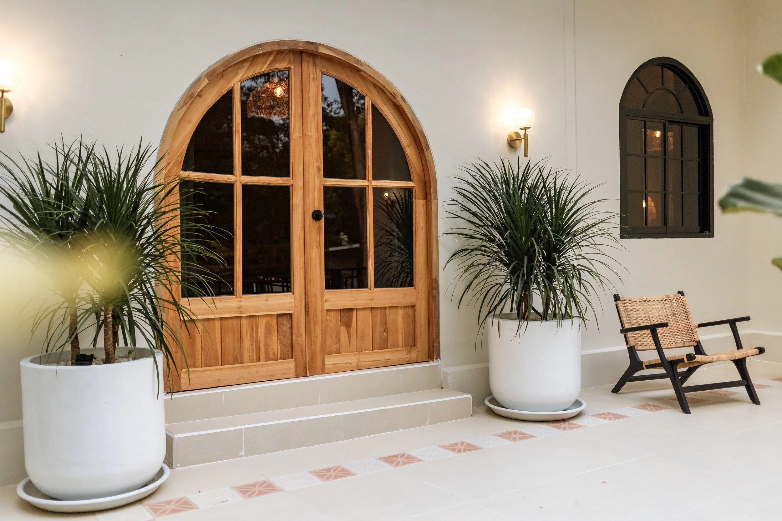 Interior view of a porch with a wooden arched door, two large potted plants, a small wooden bench, and wall-mounted lights.