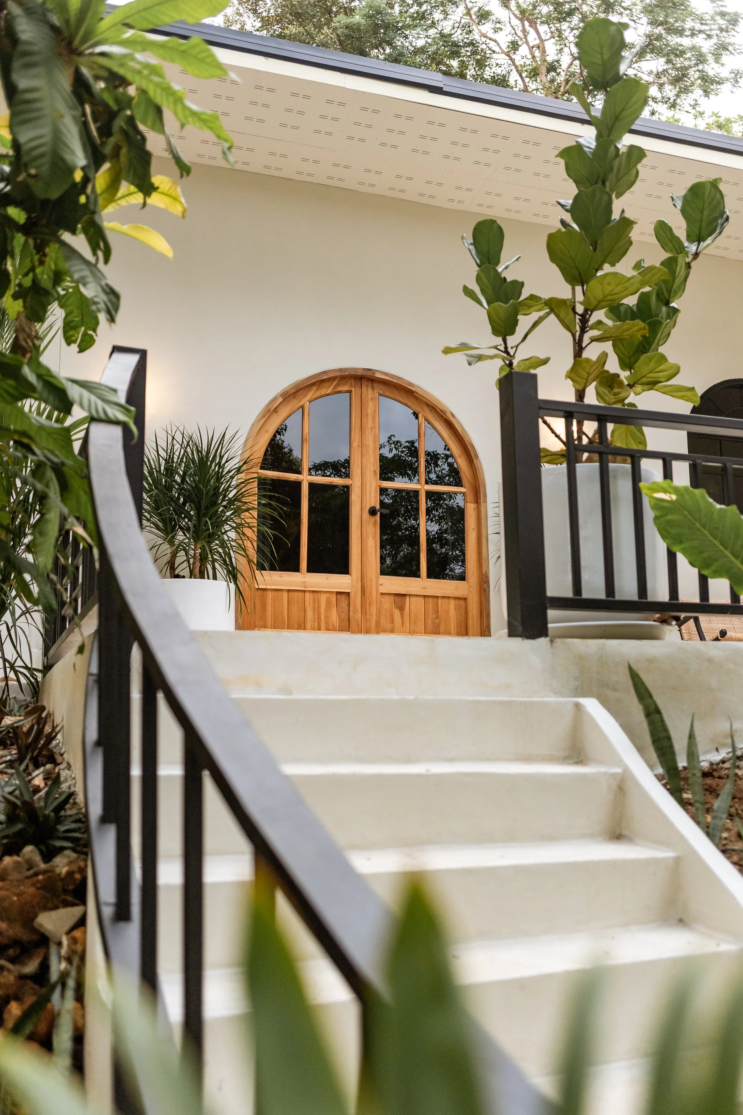 A white house with a curved wooden double door window surrounded by plants and greenery, with a black metal railing and stairs leading up to the entrance.
