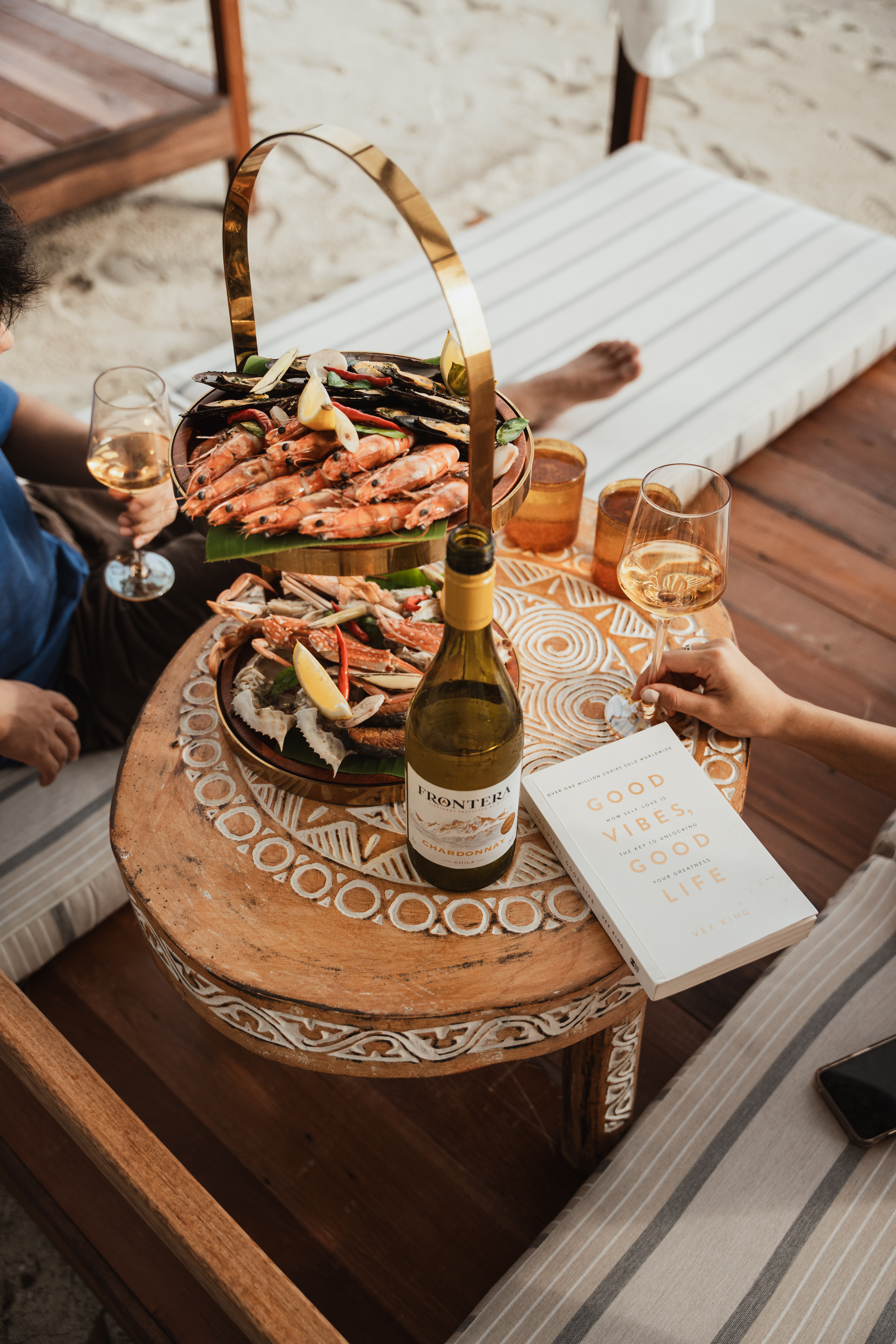 A round wooden table with a decorative carved surface, holding a bottle of wine, a book titled 'Good Vibes, Good Life,' and a two-tier seafood platter with shrimp, crab, and lemon wedges, excluding the ocean and beach setting, with two glasses of wine and two people seated nearby.