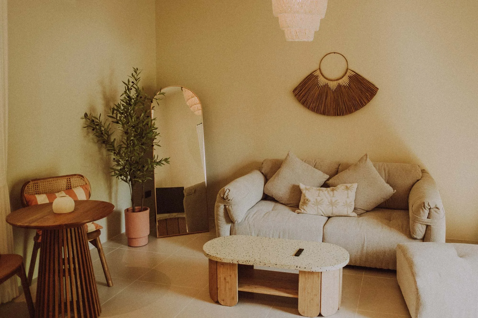 A cozy living room with beige walls and furnishings, including a cream-colored sofa with pillows, a small wooden side table, a standing mirror, a tall plant in a pink pot, and wall decor made of natural materials.