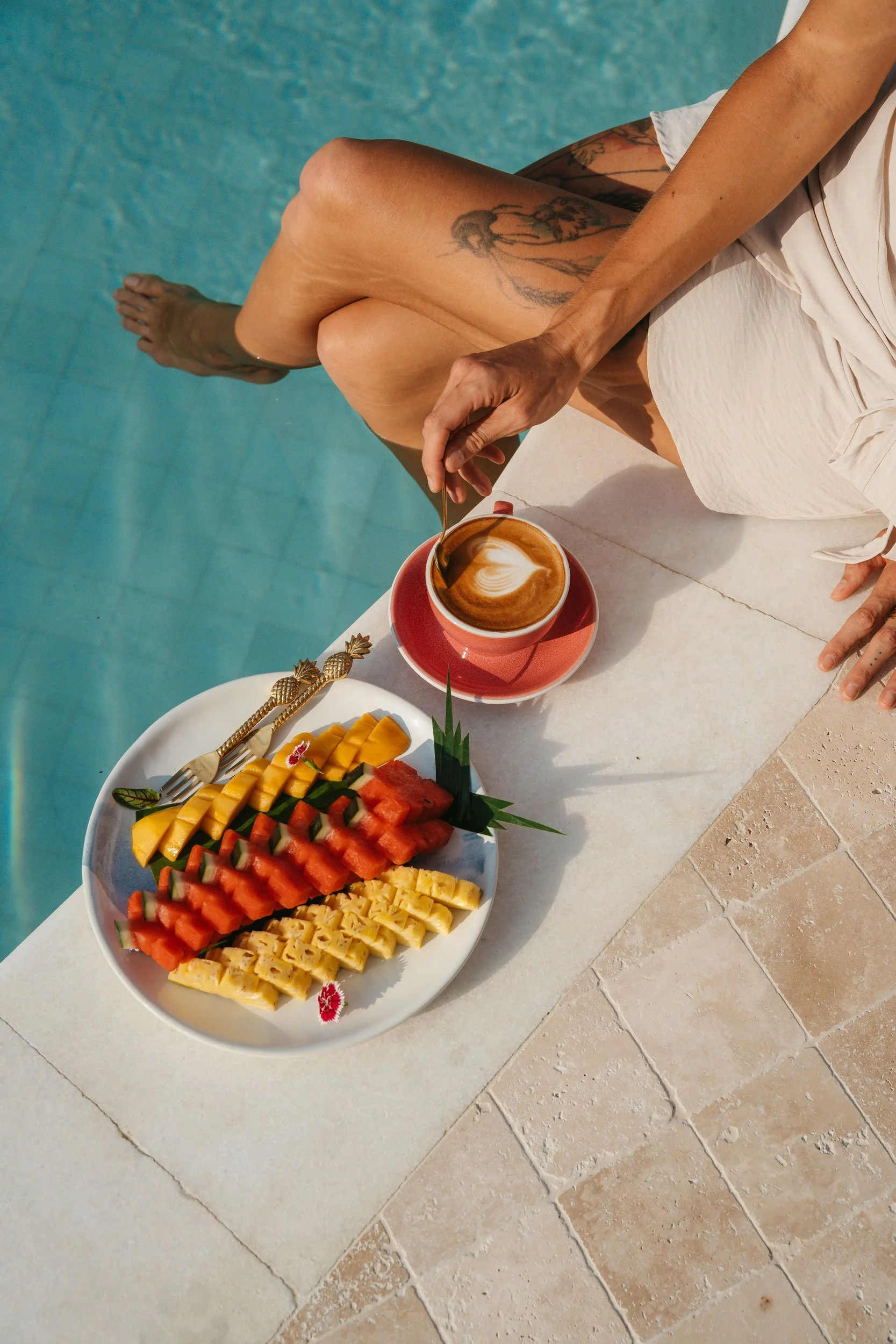 Person sitting by a pool with a plate of sliced tropical fruits and a cup of coffee.