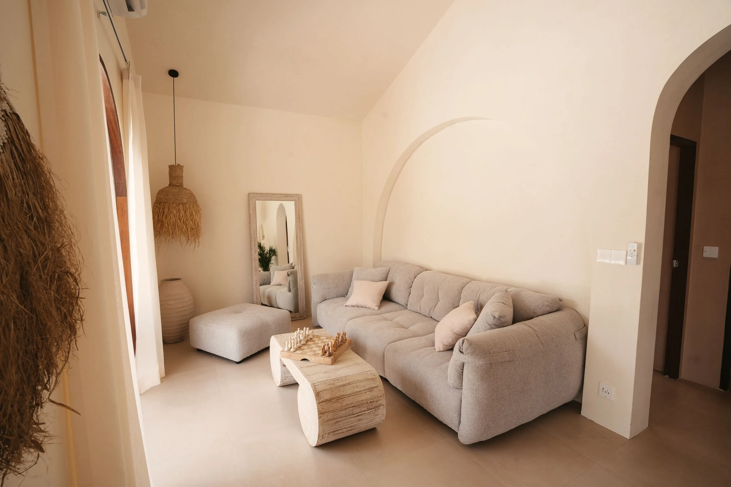 Living room with a beige couch, a tall mirror, a textured ottoman, a wooden chessboard on a curved wooden coffee table, a floor lamp with a woven shade, and neutral-colored decor.