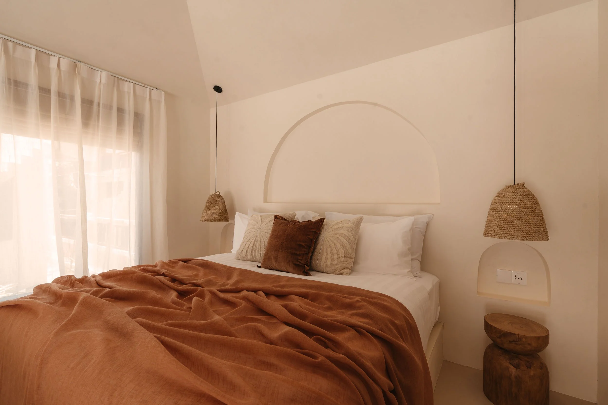 A neatly made bed with white sheets, brown velvet pillows, and a rust-colored bedspread in a minimalist bedroom with hanging wicker pendant lights and sheer white curtains.