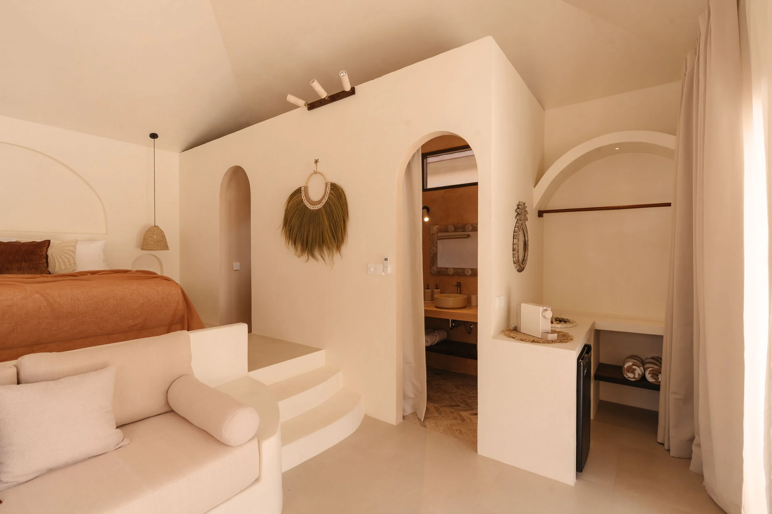 Living space with white walls, a beige sofa, a bed with a brown blanket, and a bathroom visible through an arched doorway. Decor includes a woven wall hanging and circular mirror, with curtains on the right side.