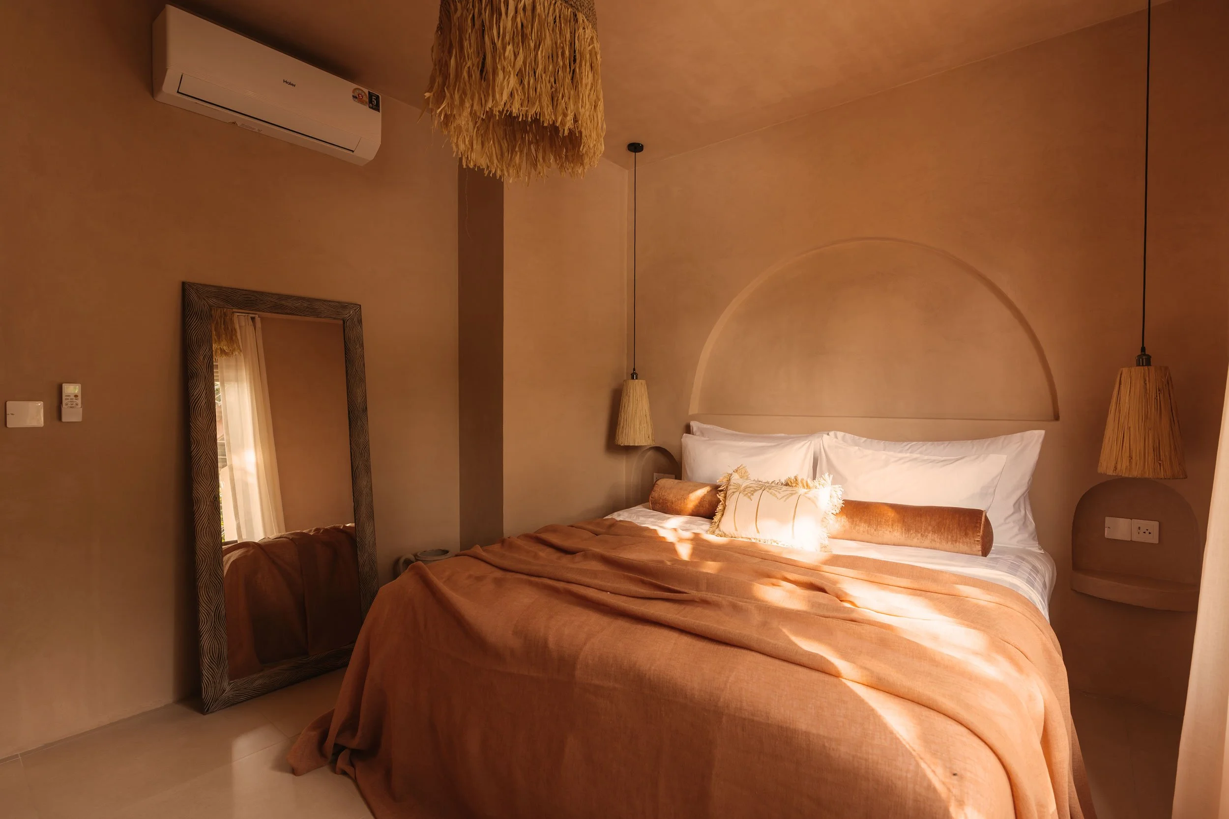 A cozy bedroom with a bed covered in beige and white linens, fluffy pillows, and warm lighting. There are hanging pendant lights on either side of the bed, a large mirror leaning against the wall, and a wall-mounted air conditioner.
