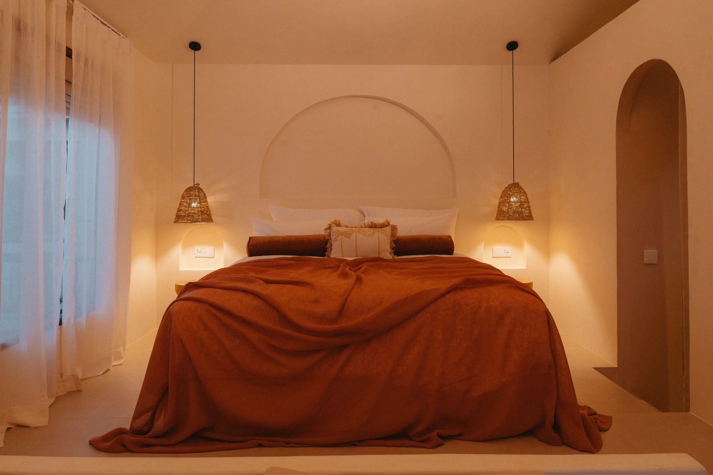 A cozy bedroom with a large bed covered in a brown blanket, two hanging pendant lights on either side, white curtains on the left, and an arched doorway on the right.