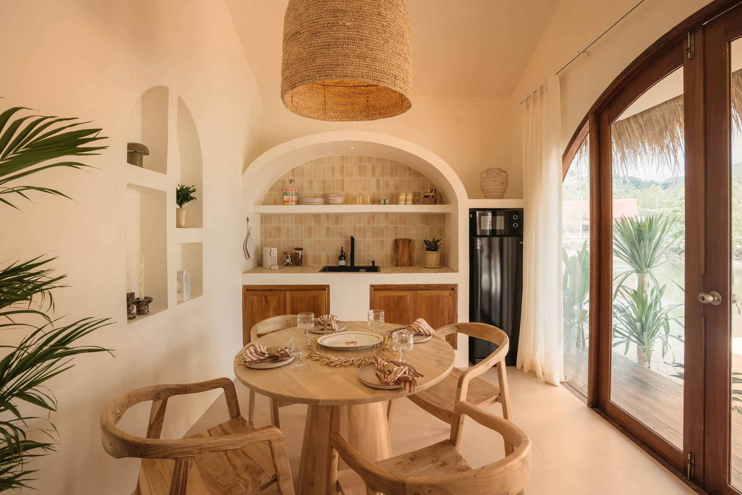 A cozy dining area with a round wooden table set with plates, glasses, and napkins. In the background, there is a small kitchenette with beige tiled backsplash, a sink, and wooden cabinets. Large glass doors with white curtains lead outside, showing 