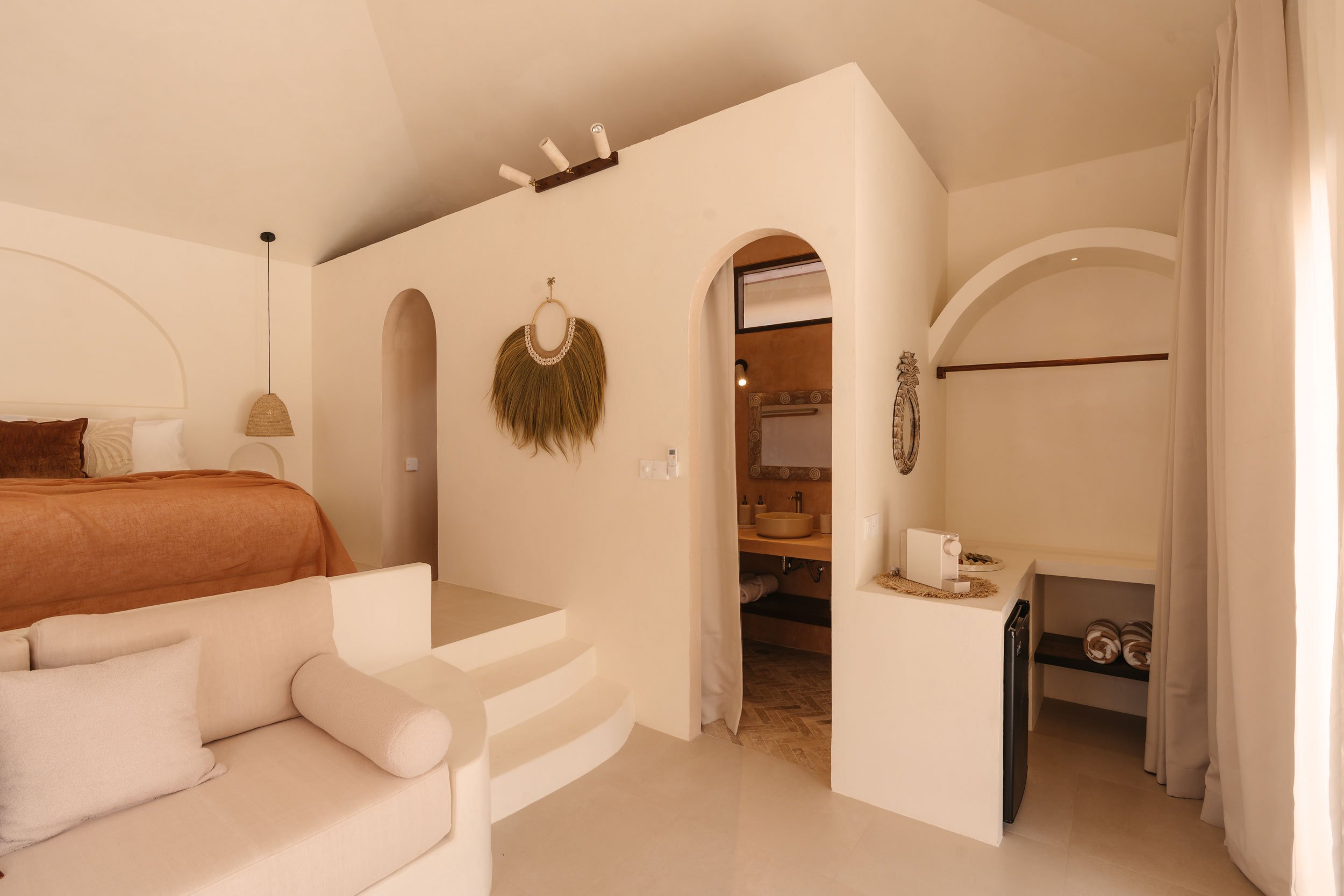 Interior of a cozy room featuring a bed with rust-colored bedding, a white couch in the foreground, artistic wall hangings, and a small bathroom visible through an arched doorway.