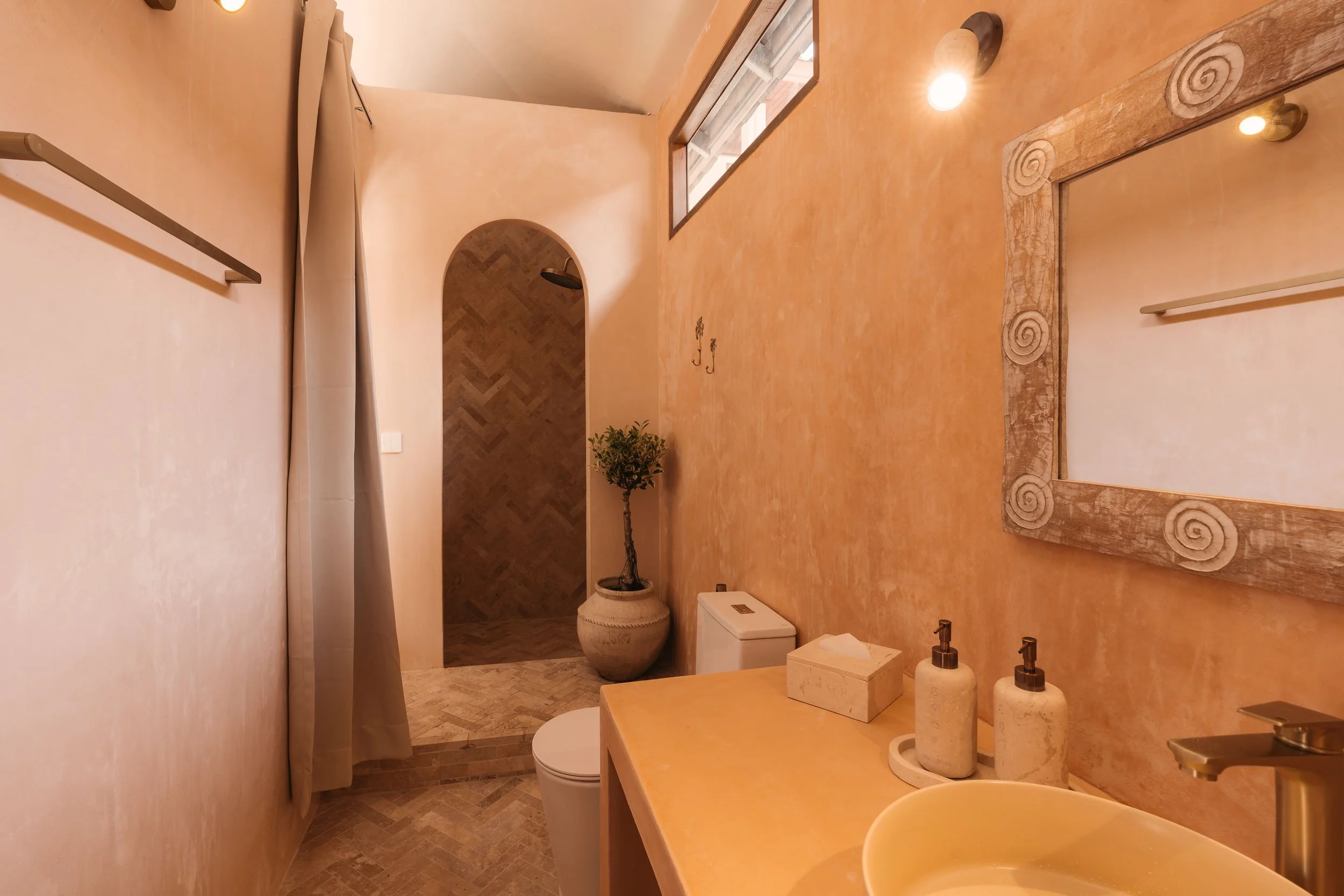 A small bathroom with peach-colored walls, a large mirror with a swirl-patterned wooden frame, two soap dispensers, a tissue box, a calendar, and a sink. There is a toilet near a potted plant, a shower area with a curtain, and a small window near the