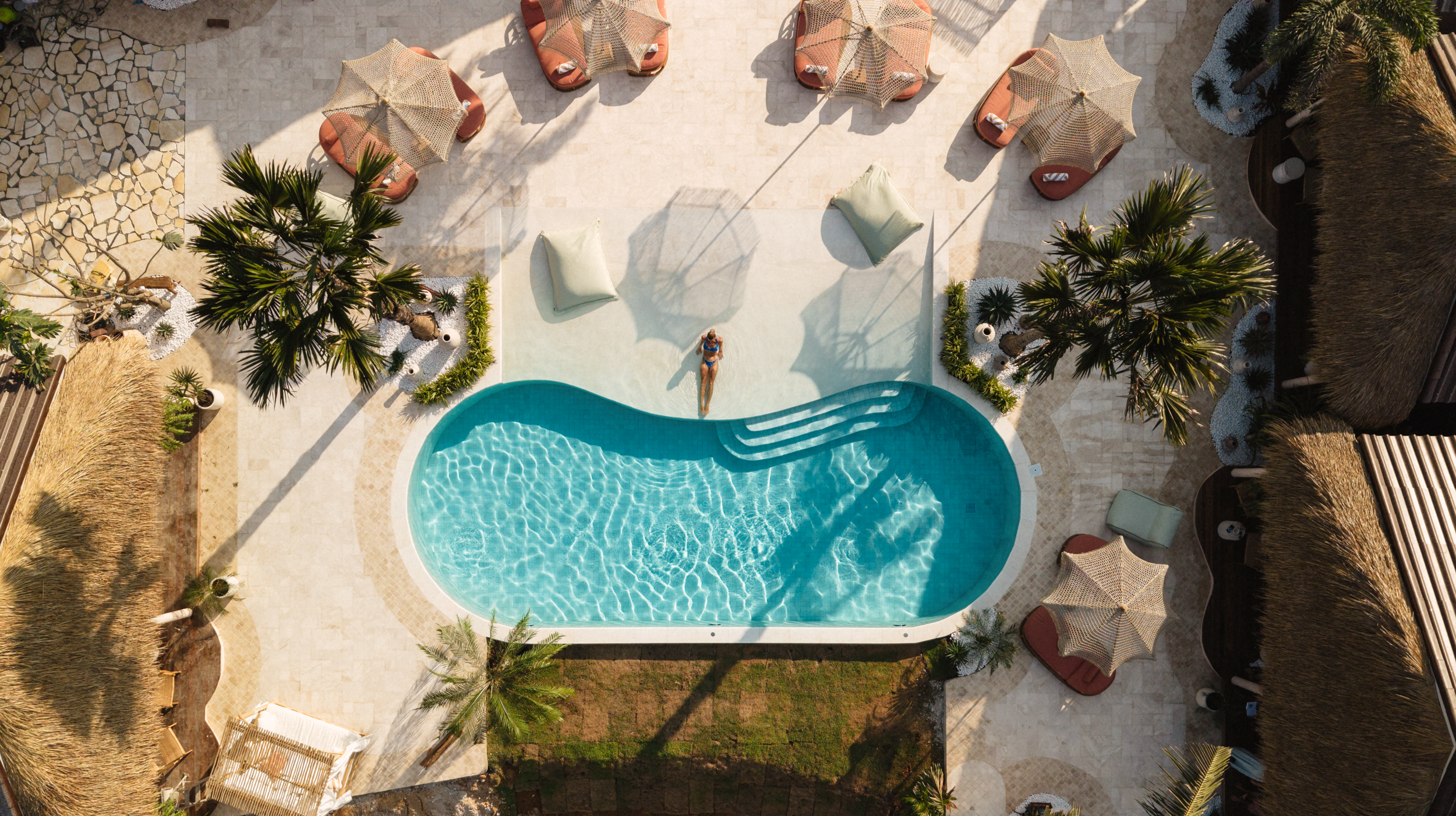 Aerial view of a hotel pool area with a woman sunbathing on a float in the pool, surrounded by palm trees, sun umbrellas, and lounge chairs.