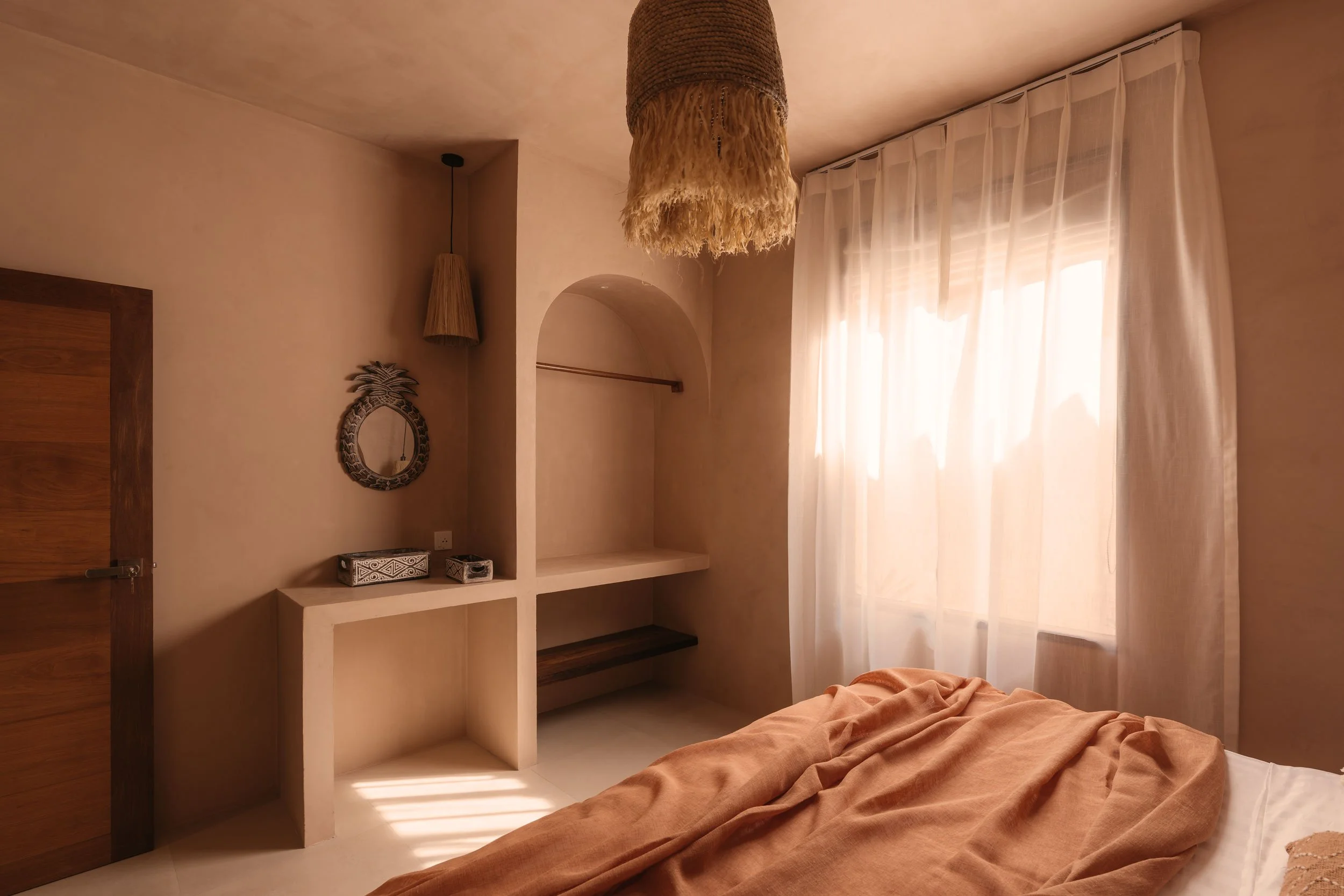 Sunlit bedroom with beige walls, sheer curtains, a bed with an orange blanket, a wooden door, a decorative pineapple-shaped mirror, and woven pendant lights hanging from the ceiling.