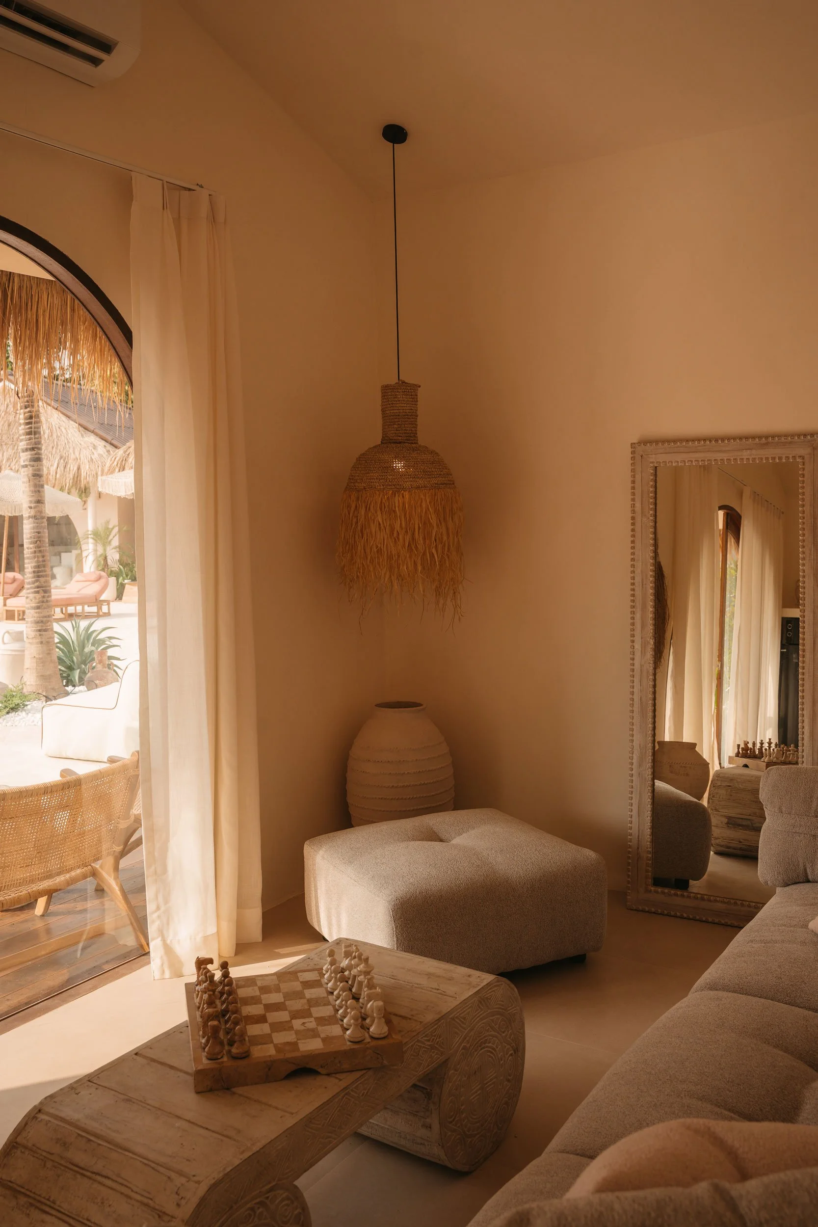 A cozy living room with beige furniture, a large mirror, chandelier, and a chessboard on a wooden coffee table, with sunlight streaming through sheer curtains onto a patio with lounge chairs and tropical plants.
