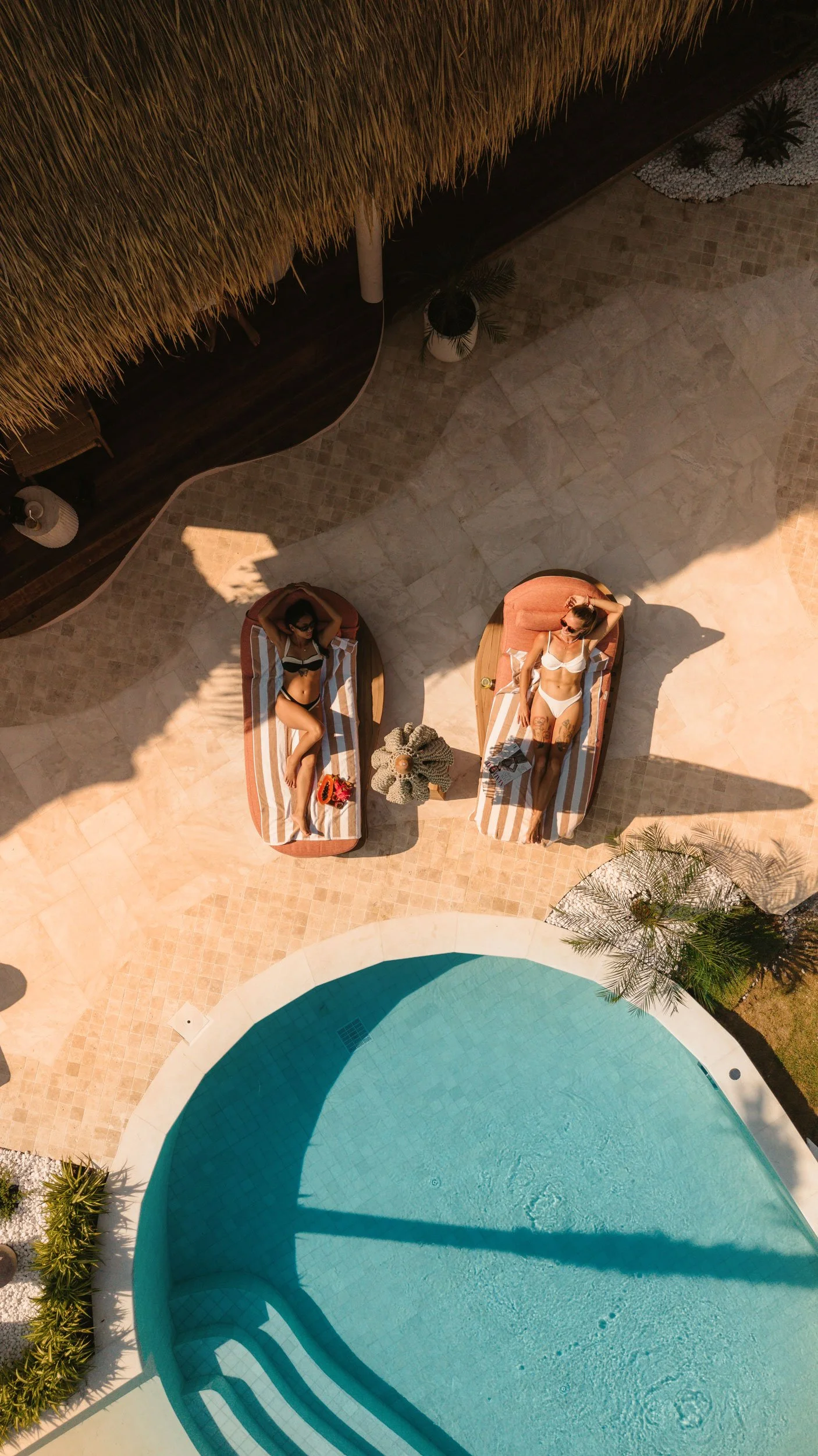 Two women sunbathing on lounge chairs by a swimming pool, with a thatched roof and plants nearby, taken from above in bright sunlight.
