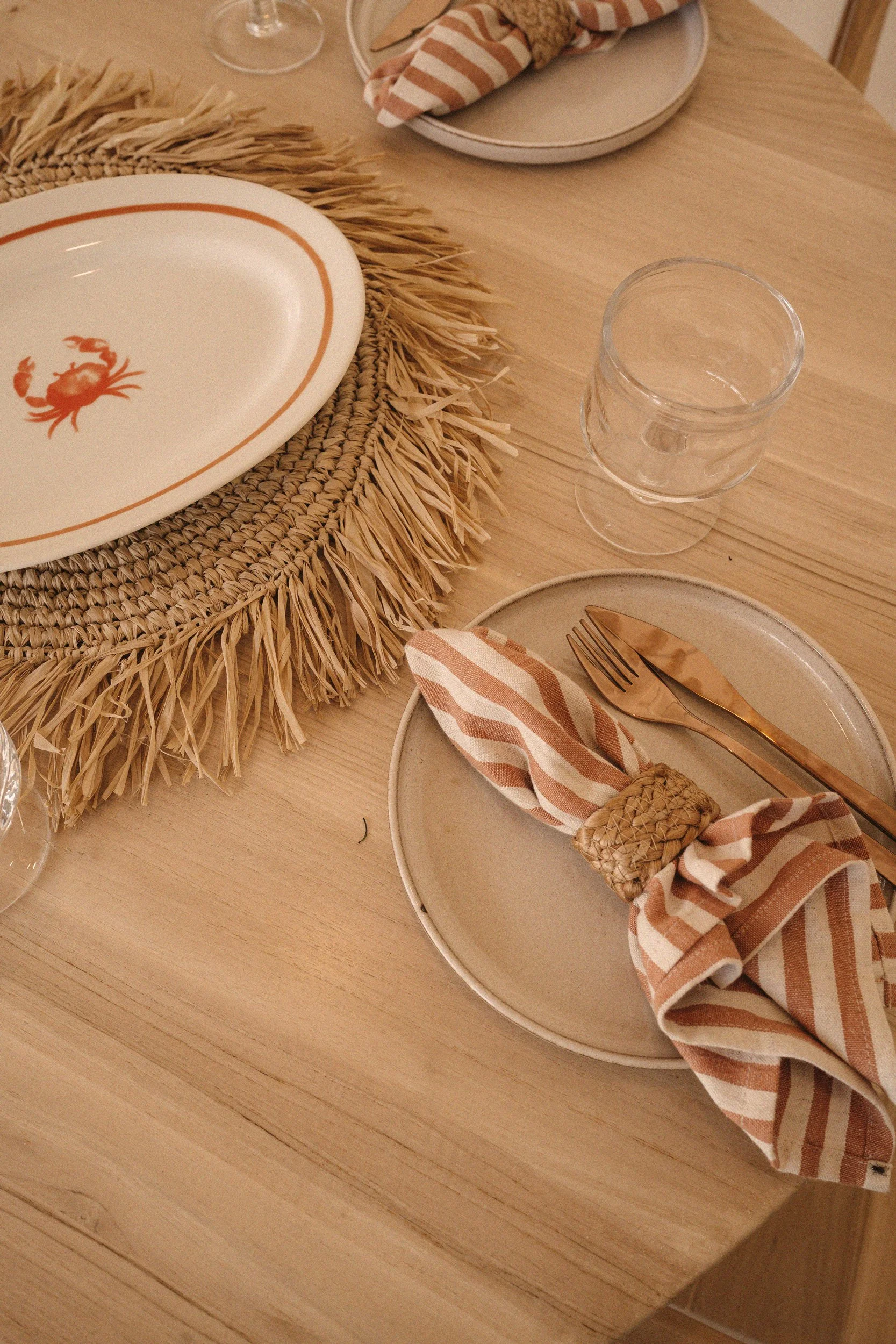 Table setting with a round woven placemat, a white plate with a crab design, a striped cloth napkin wrapped around cutlery, a clear glass cup, and additional plates and utensils in the background.