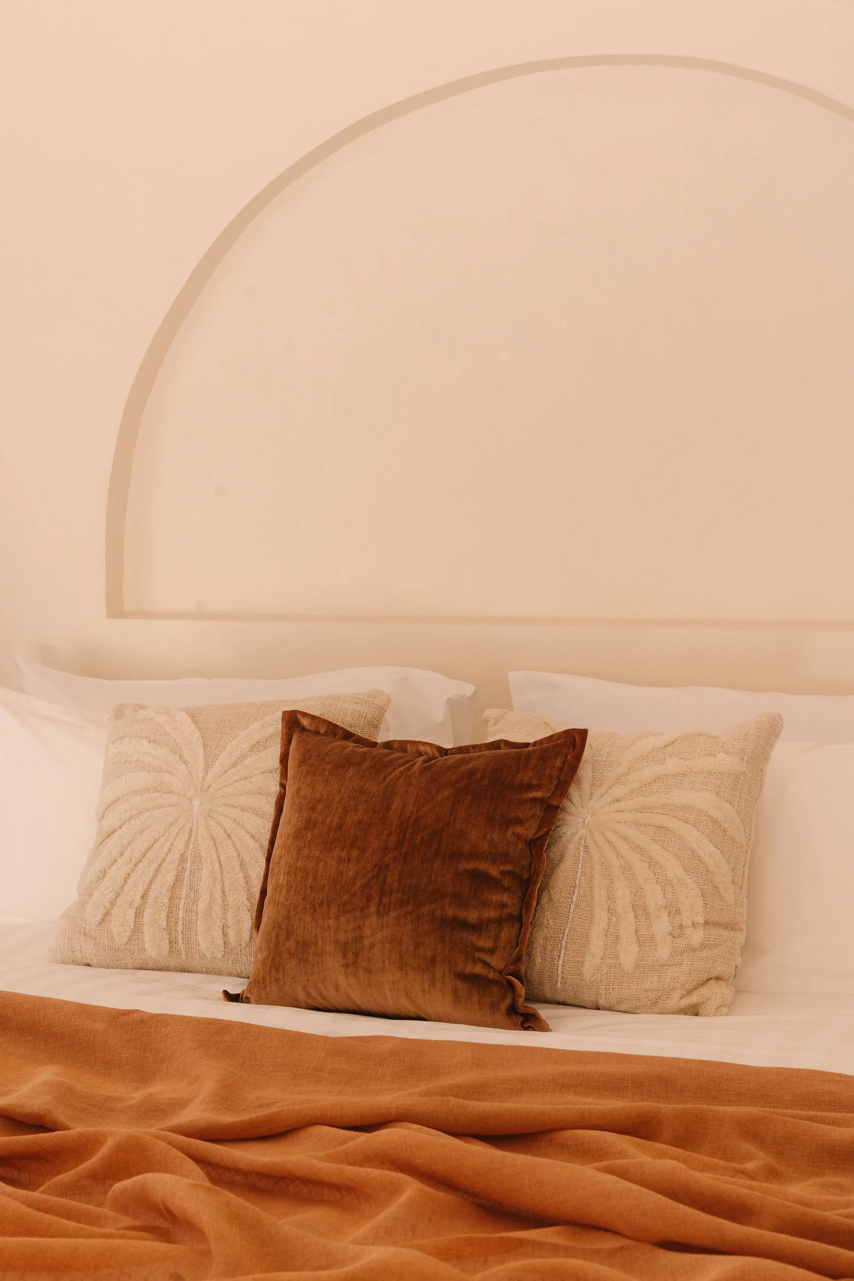 A neatly made bed with a beige headboard, white pillows, embroidered beige pillows, and a brown velvet accent pillow. The bed has warm-toned bedding.