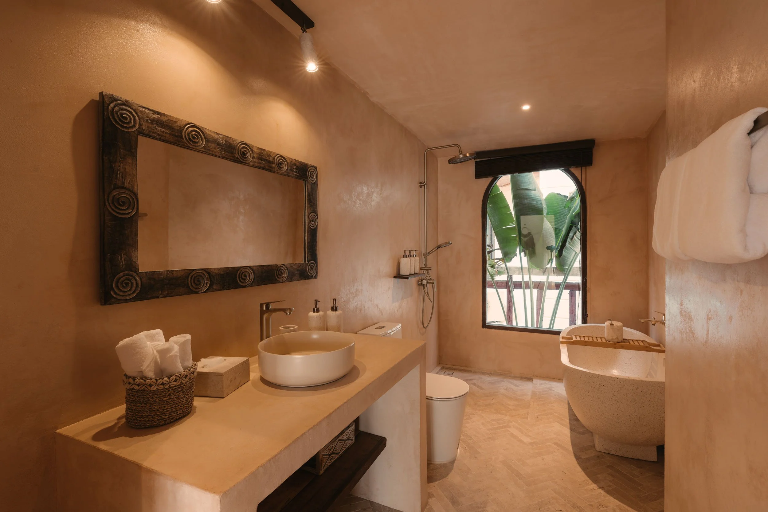 Modern bathroom with beige textured walls, large mirror with decorative frame, vessel sink, soap dispensers, toilet, bathtub, and window overlooking tropical plants.