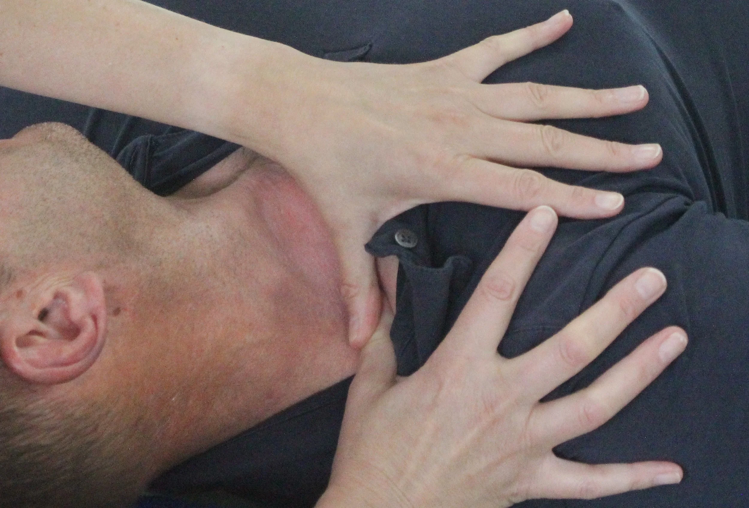 A person with a hand resting on their chest, lying down on a dark surface, wearing a dark shirt, with part of their face visible including an ear and hair.
