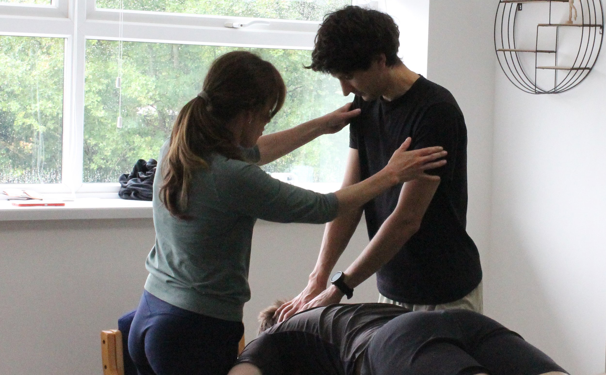 A woman with long hair assists a man with curly hair during a medical neck or spine injury examination, in a room with large window and greenery outside.