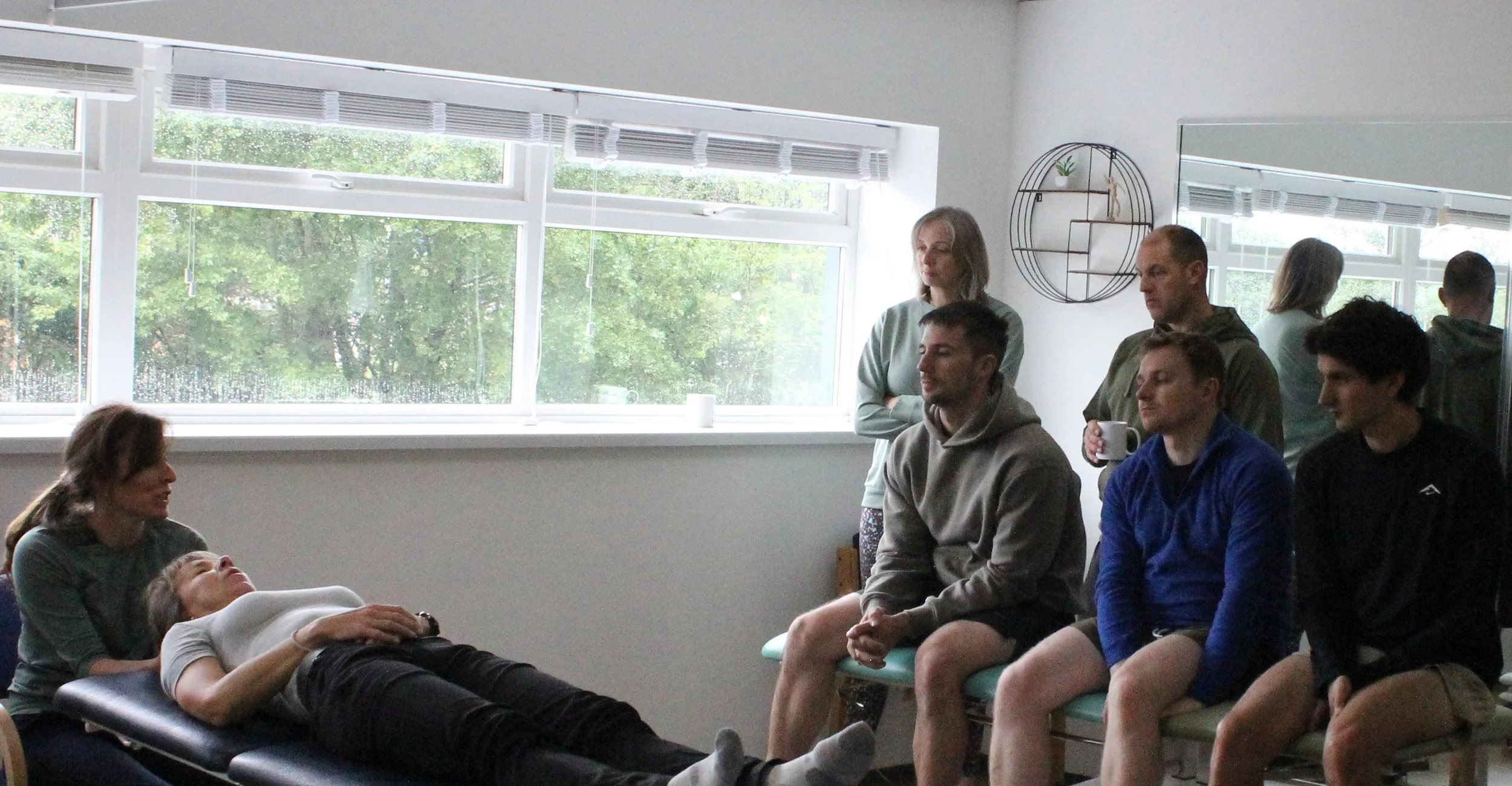 A group of people attending a first aid training or CPR class in a room with large windows, some sitting on chairs and one person lying on a training mannequin.