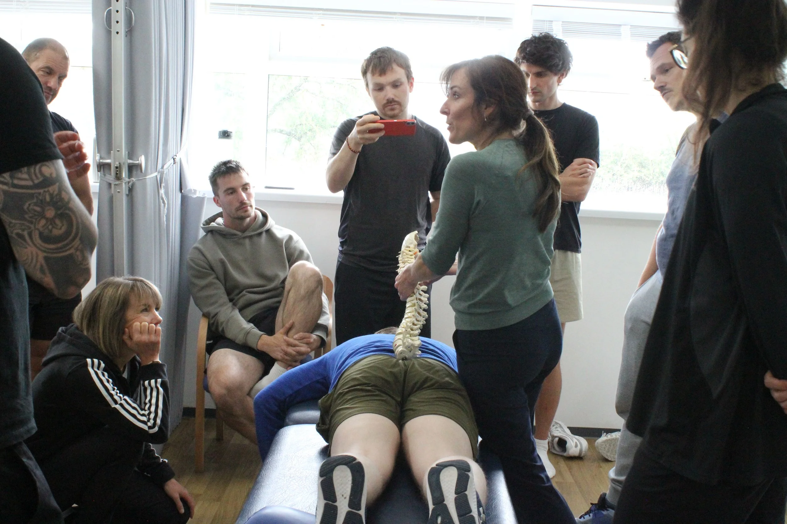 A group of people gathered around a person lying face down on a table, with a skeleton model held by a woman, implying a medical or educational demonstration.