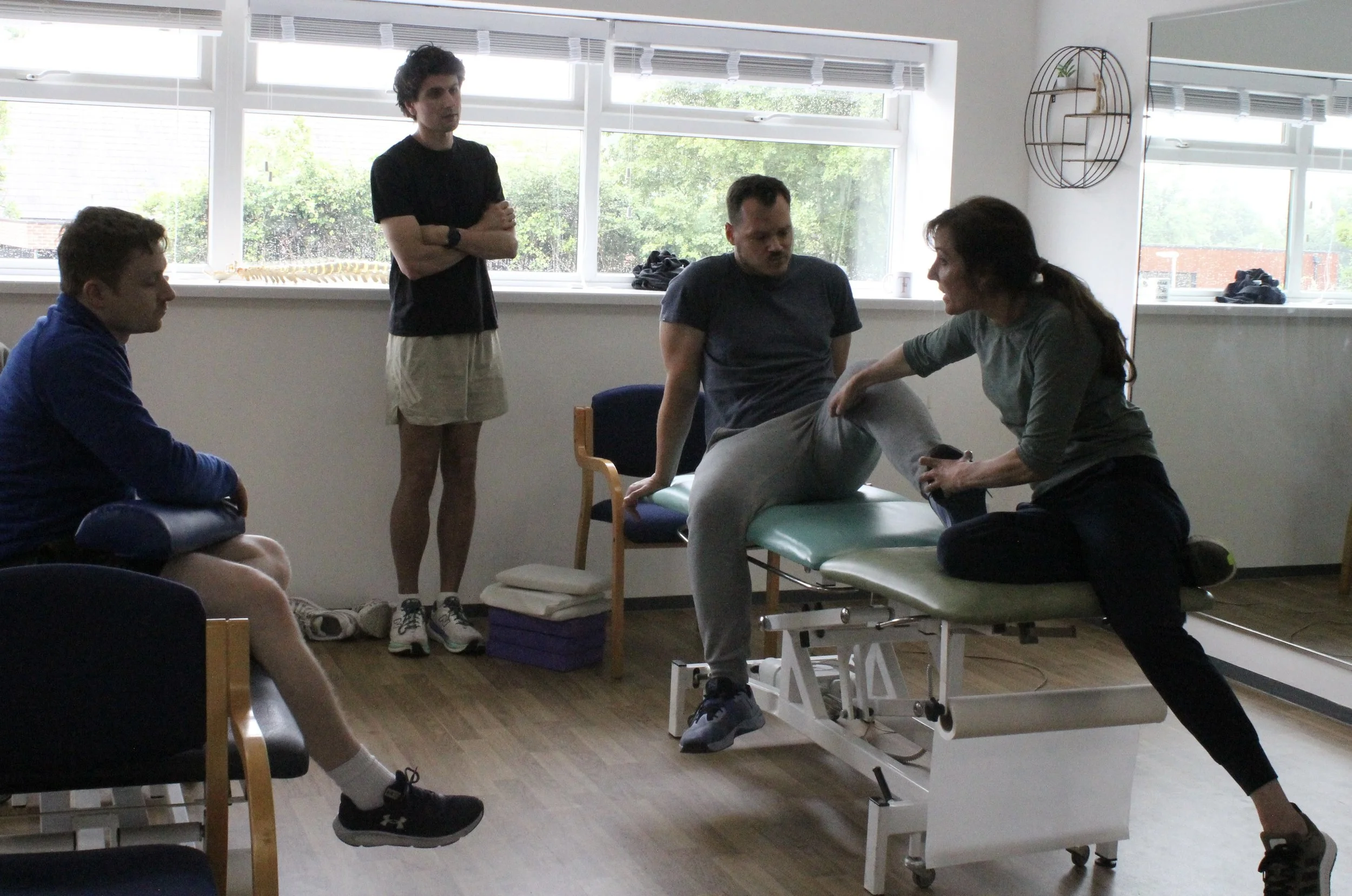 A group of five people, three men and two women, in a room with large windows, participating in a physical therapy or rehabilitation session. One woman is assisting a man on an examination table, while others observe.