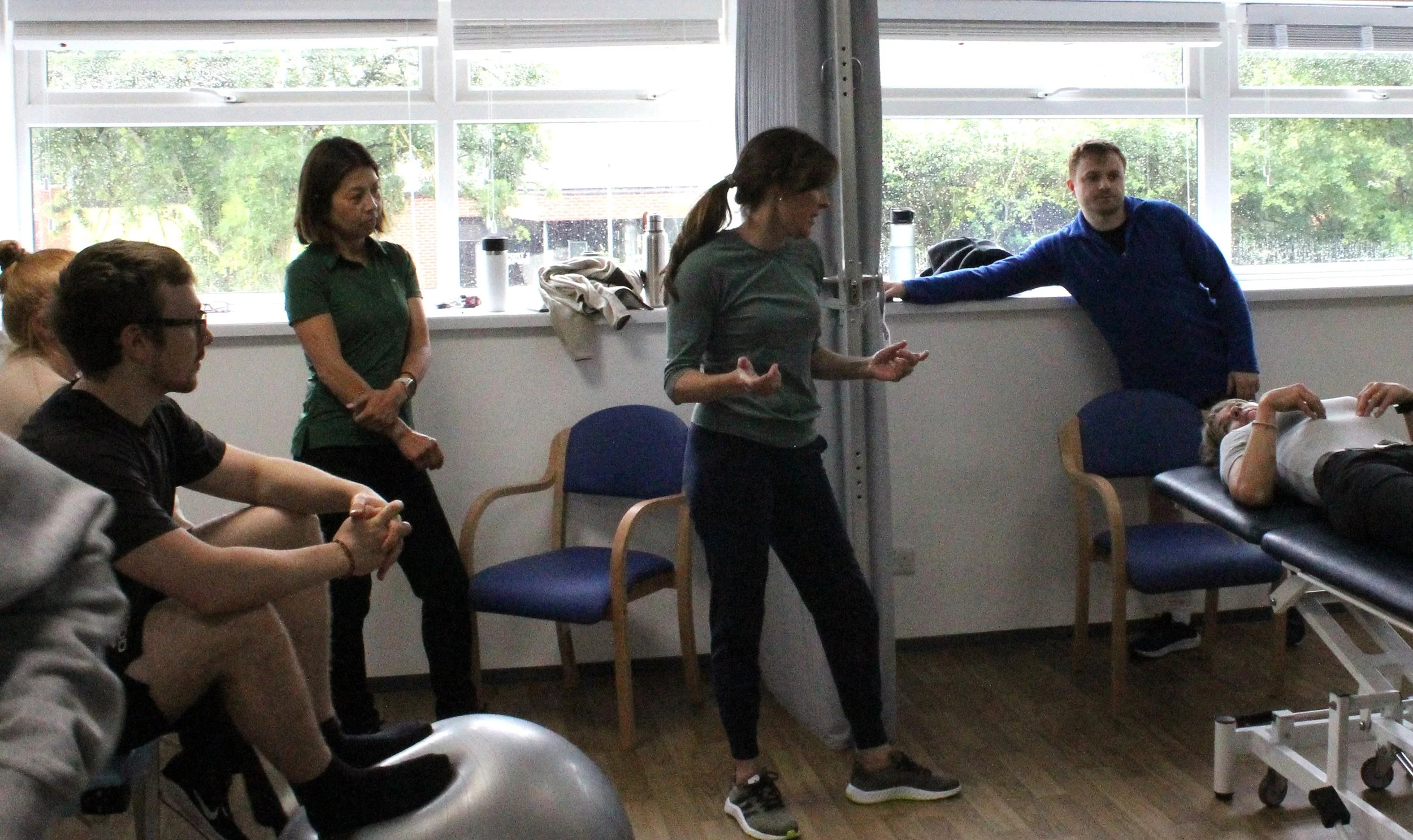 A group of people in a medical or physical therapy setting. Some are seated, and one woman is standing and talking. Another woman is lying on a medical examination table, with a man leaning on the wall nearby. The room has large windows showing outdoor greenery.