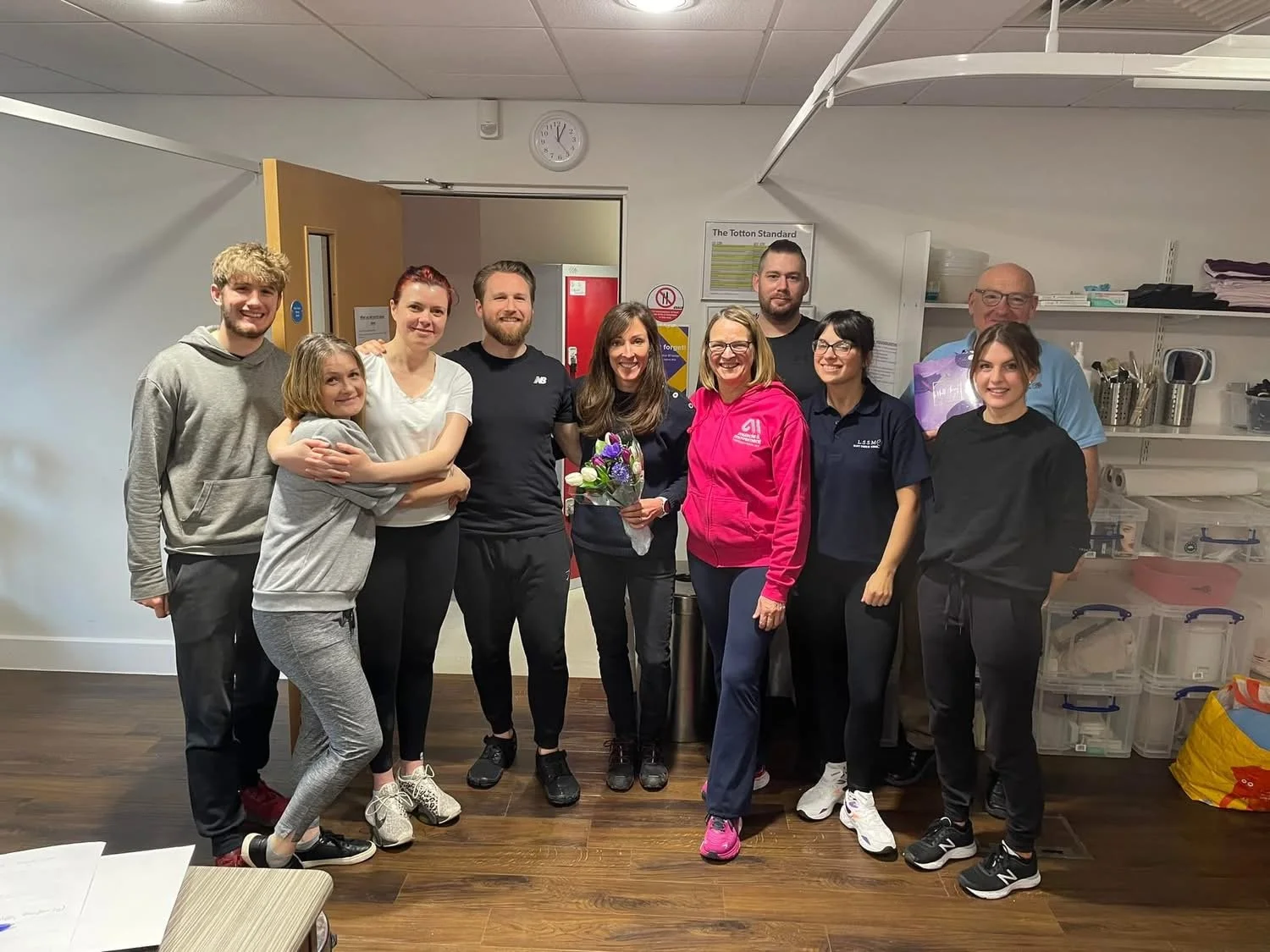 Group of eleven people standing inside a room, some smiling, with three women in the center holding flowers, and a kitchen or storage area with shelves and containers in the background.