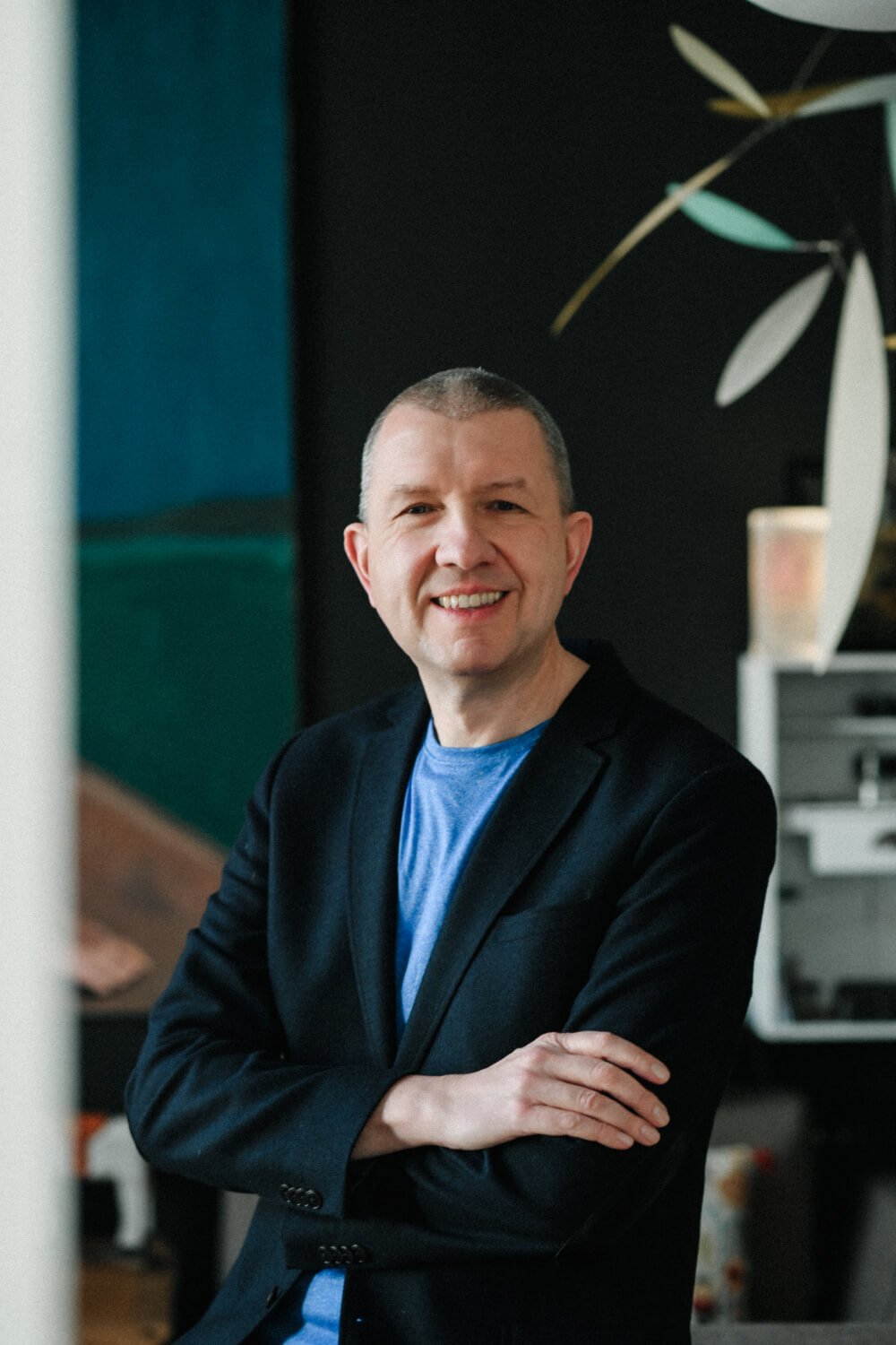 Ein lachender Mann mit Kurzhaarfrisur in dunklem Blazer und blauer T-Shirt sitzt in einem Raum mit dunklem Hintergrund und Pflanzen.