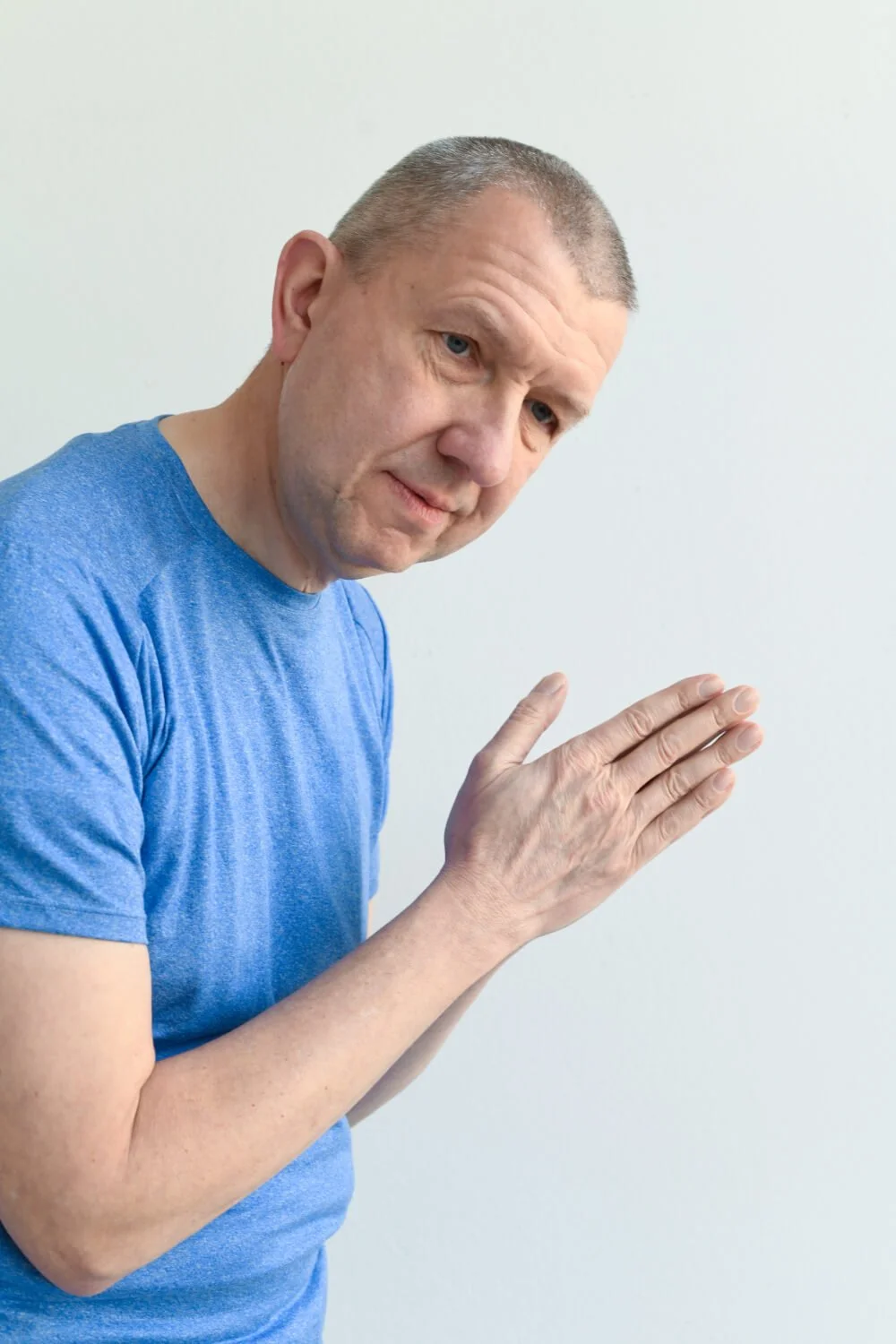 Ein älterer Mann trägt ein blaues T-Shirt und hält seine rechte Hand in der Geste des Betens oder der asiatischen Begrüßung vor sich, vor einem weißen Hintergrund.