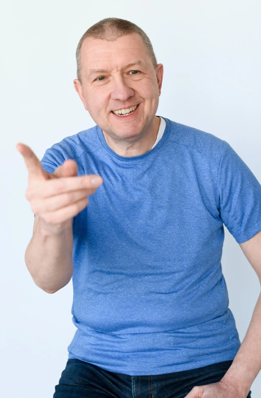 Lächelnder Mann in blauer T-Shirt zeigt nach vorne und sitzt vor weißem Hintergrund.