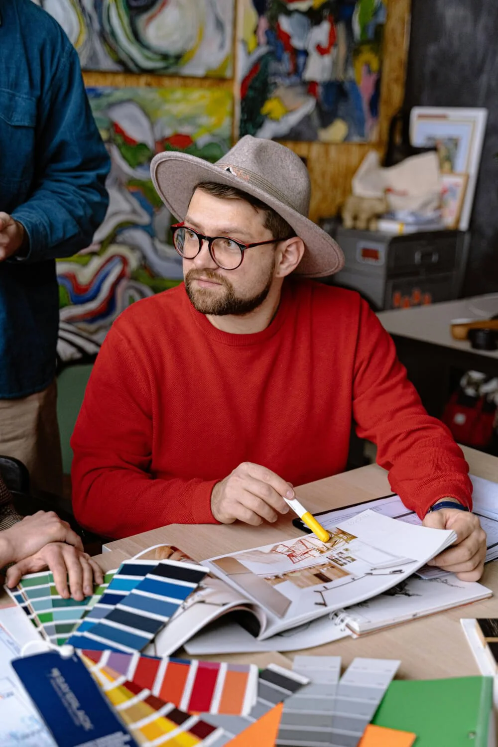 Mann in rotem Pullover, mit Brille und Hut, sitzt an einem Tisch mit Farbmustern und Gestaltungsmagazinen, während im Hintergrund bunte Gemälde und Kunstwerke zu sehen sind.