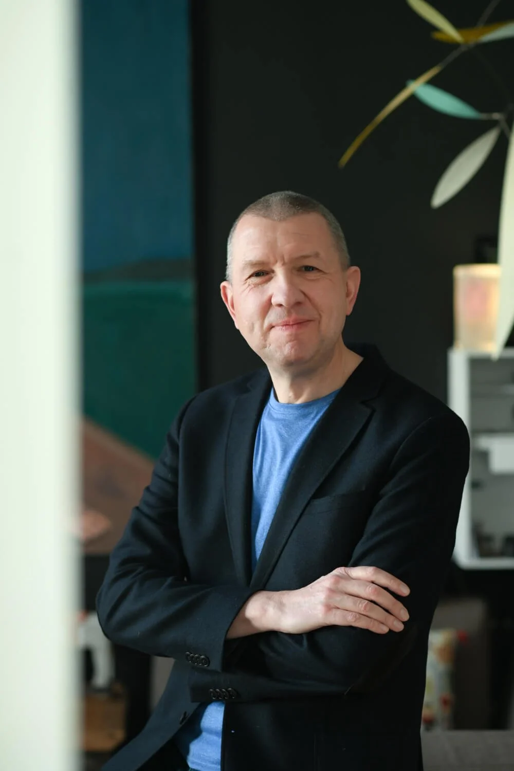 Ein Mann in dunklem Jacke und blauer T-Shirt steht in einem Raum mit dunklem Hintergrund und schaut in die Kamera.