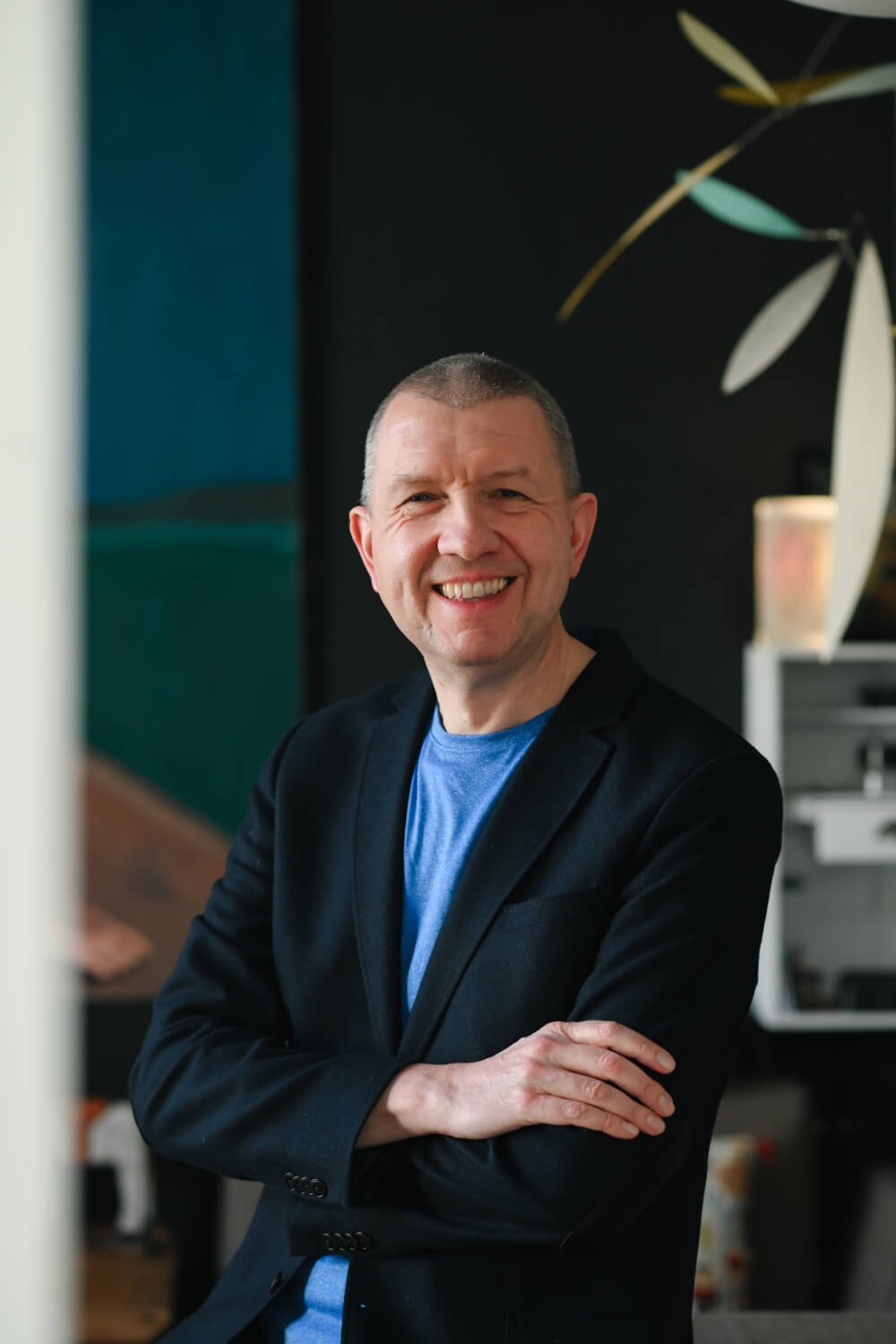 Ein lachender Mann mit kurzhaarigem Haar in einem schwarzen Blazer und bläulichem T-Shirt, in einem modernen Raum mit dunklem Hintergrund und Pflanzen.