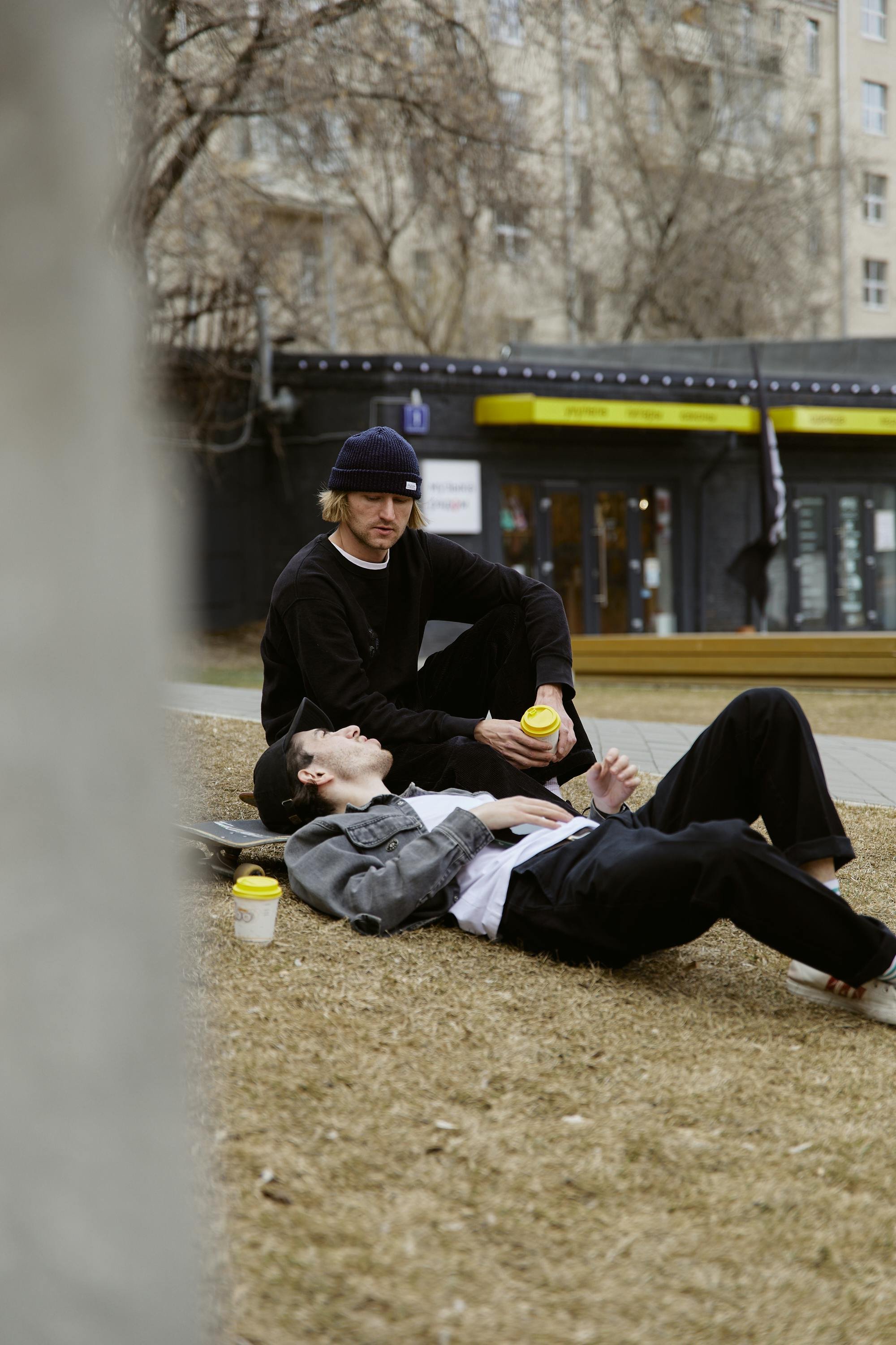 Zwei junge Männer entspannen sich auf einer Wiese im Park, einer liegt auf dem Rücken und der andere sitzt neben ihm, beide halten To-Go-Kaffeebecher. Hinter ihnen sind Bäume und ein Gebäude sichtbar.