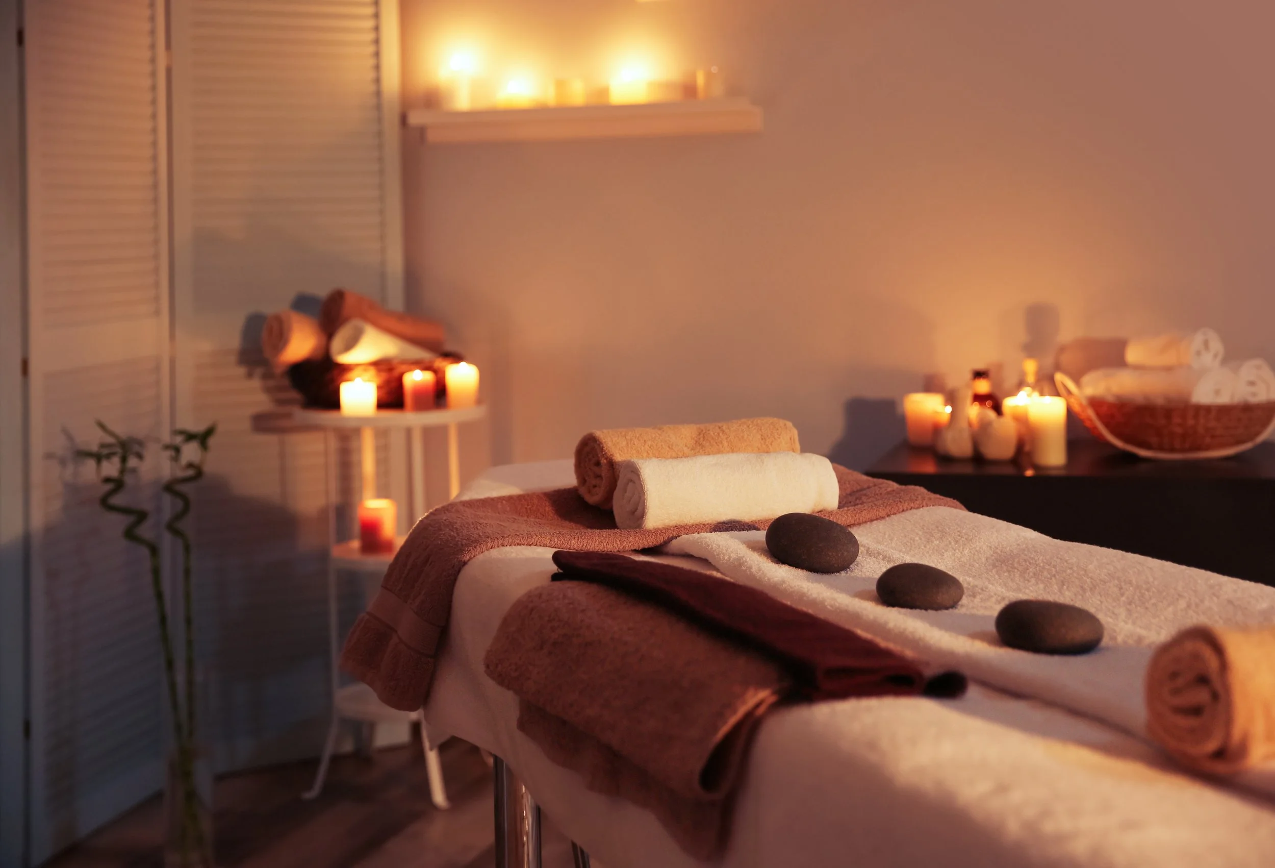 Spa massage room with candles, rolled towels, and massage stones arranged on the bed and nearby surfaces, creating a relaxing atmosphere.