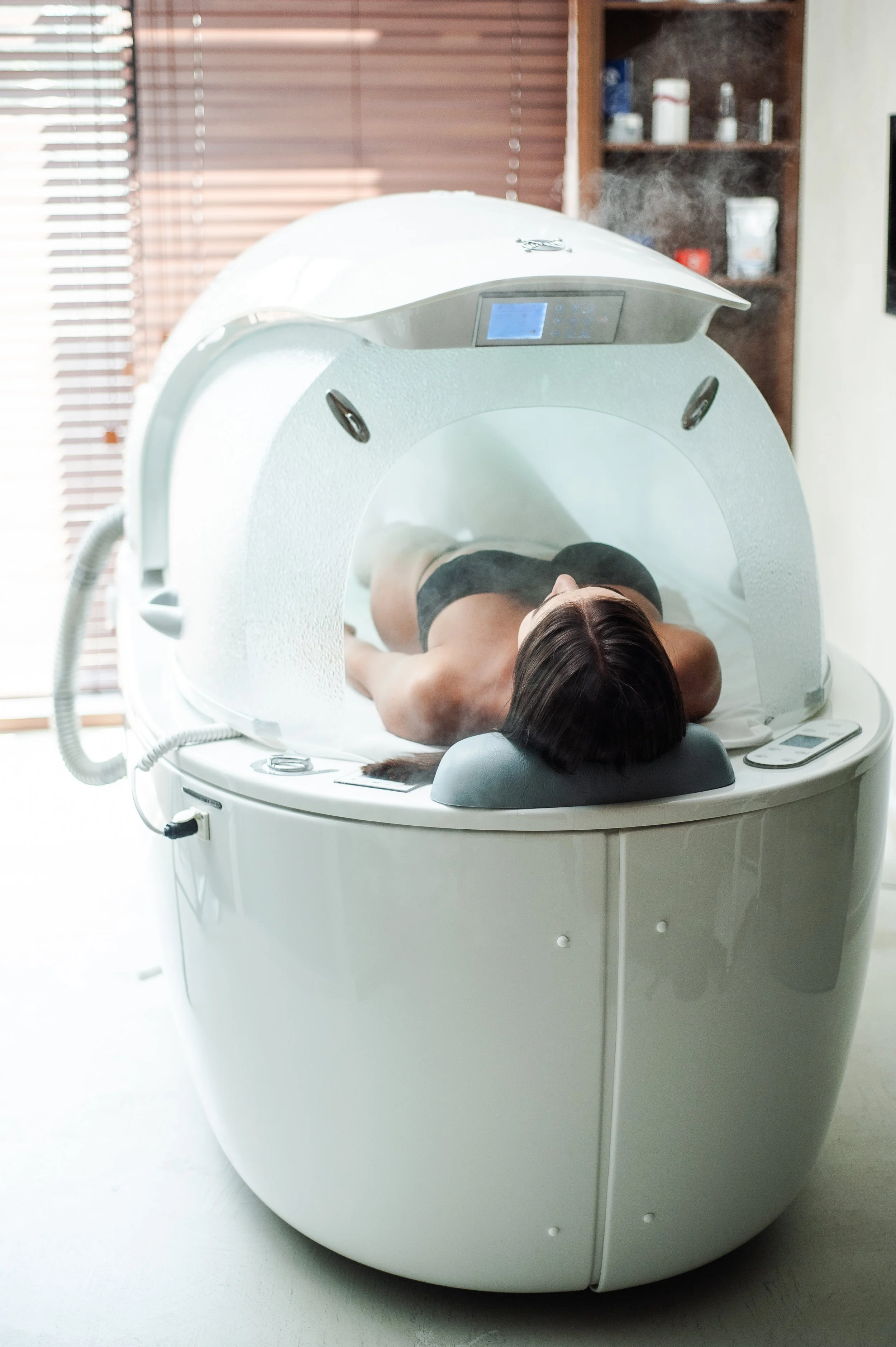 Person lying inside a white, enclosed medical machine, likely an MRI scanner, in a room with wooden blinds.