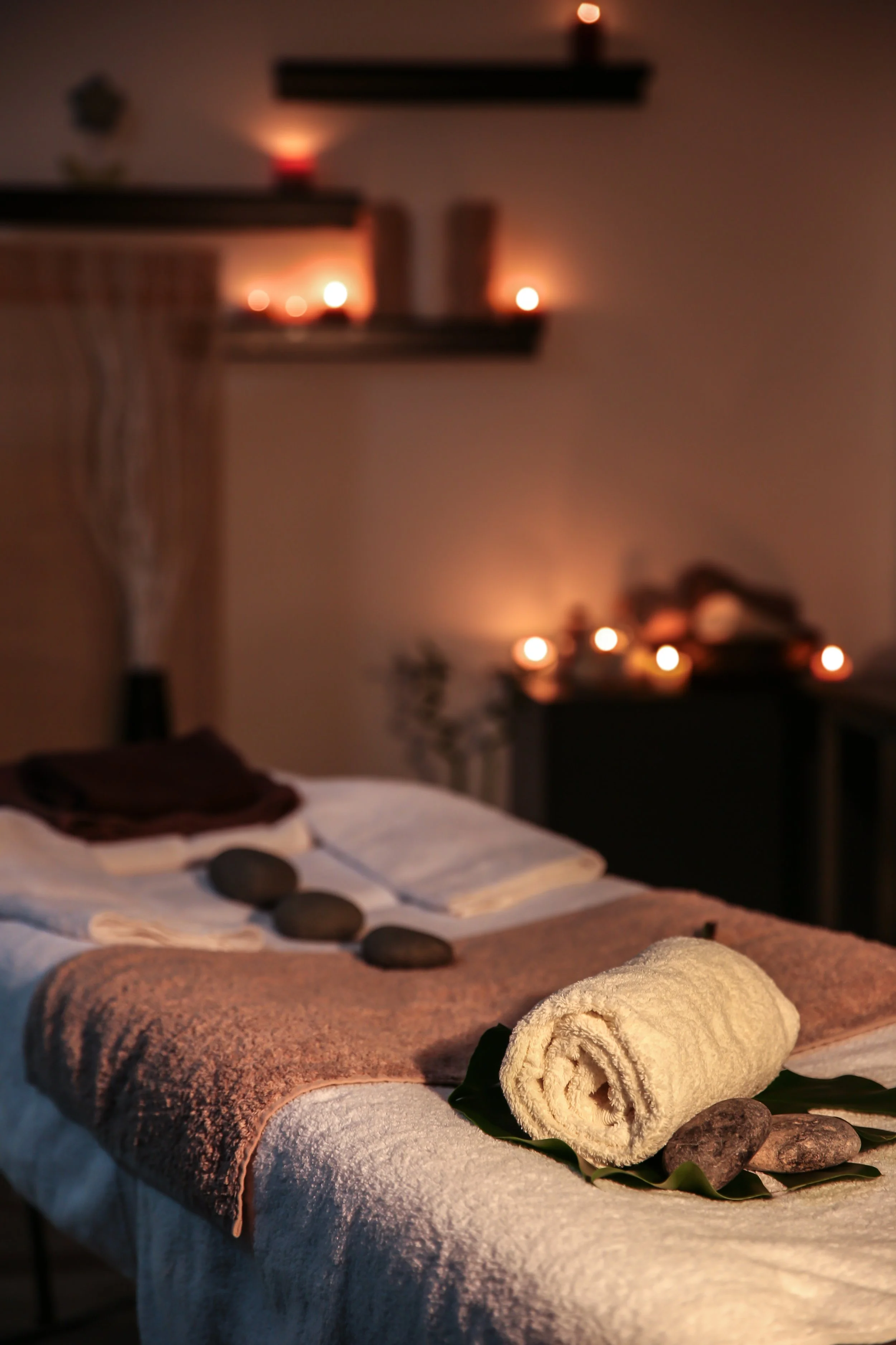 A massage table with a rolled towel and stones on it, in a dimly lit spa room with candles and soft lighting.