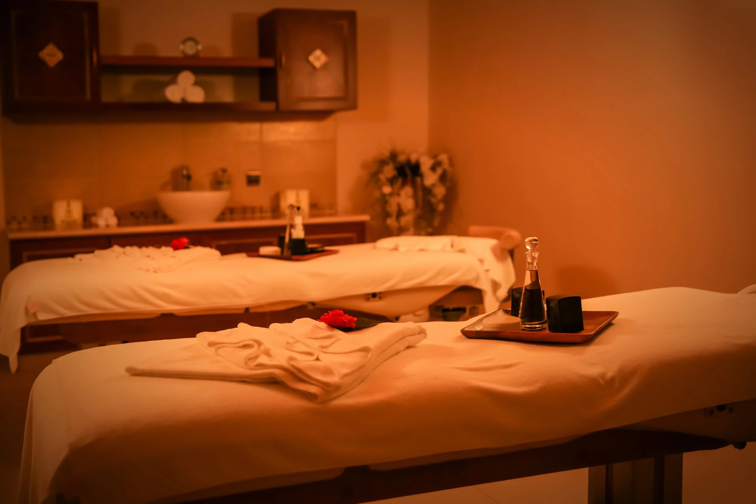 Spa room with two massage beds, each with a folded towel and a red flower, dimly lit for relaxation.