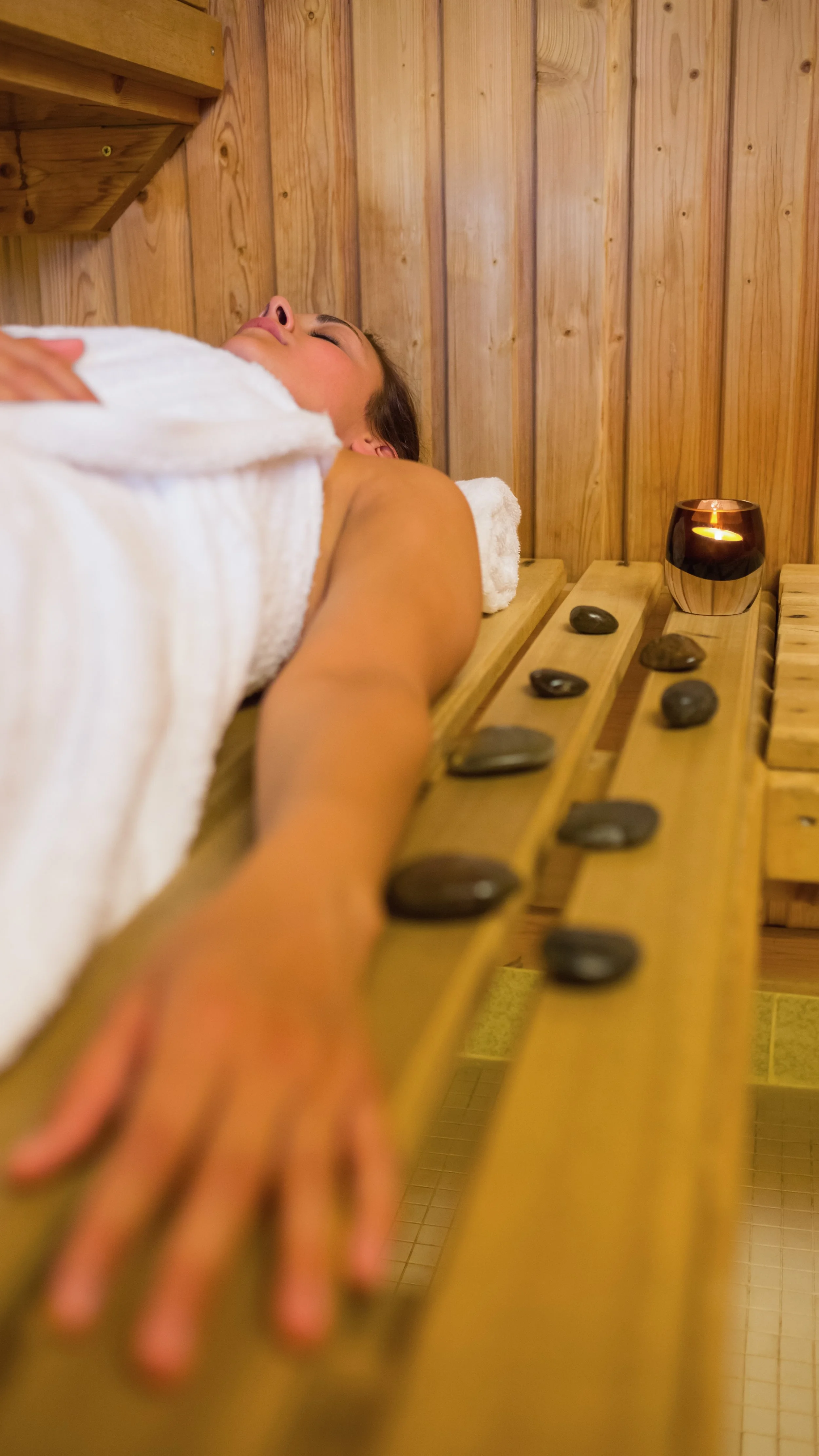A woman lying on a wooden massage table during a hot stone massage, with smooth black stones placed along her arm and a lit candle nearby in a sauna-like setting.