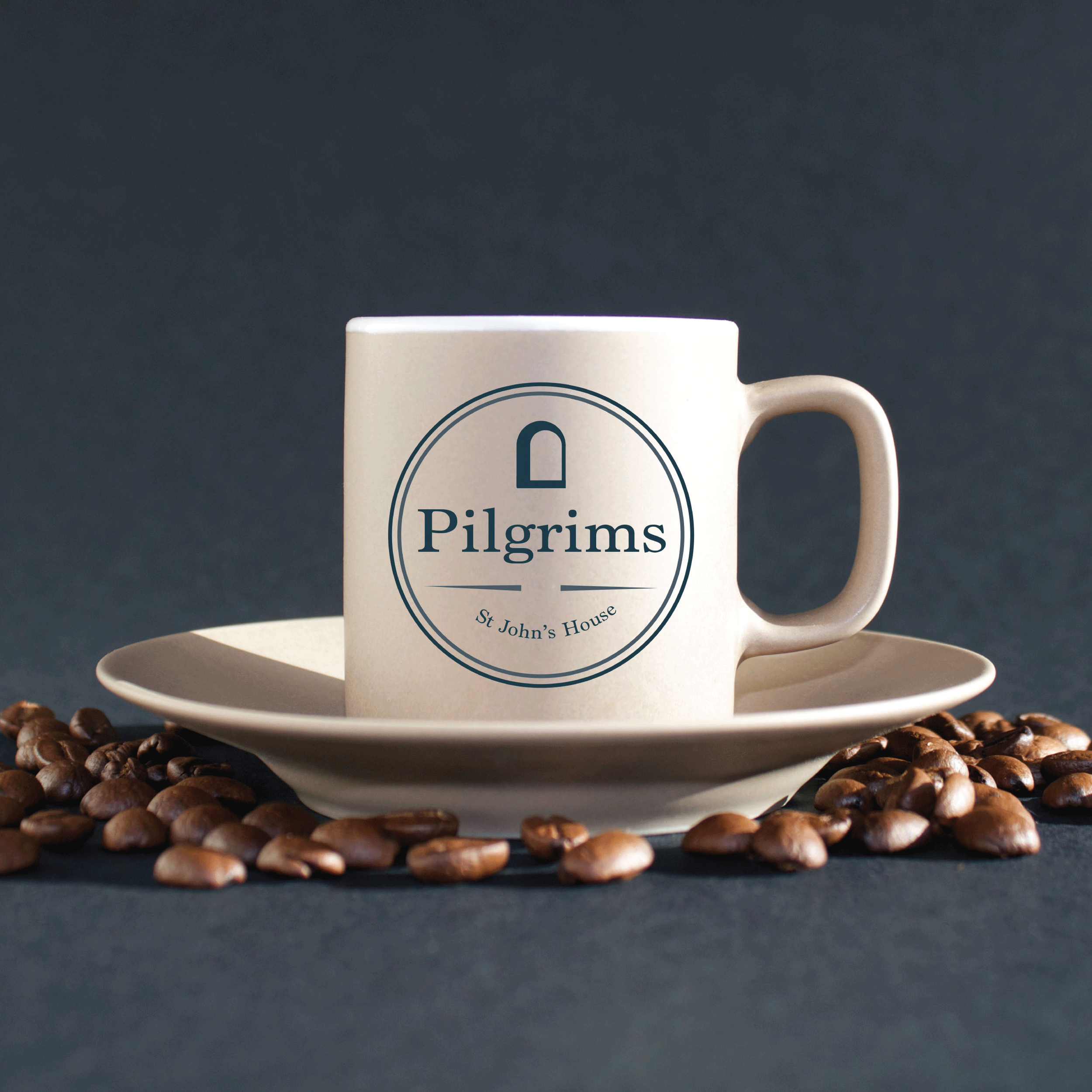 A white coffee mug on a cream saucer, decorated with a circular emblem reading "Pilgrims," with a small door icon at the top and the words "St John’s House" underneath. Coffee beans are scattered around the saucer, set against a dark background.