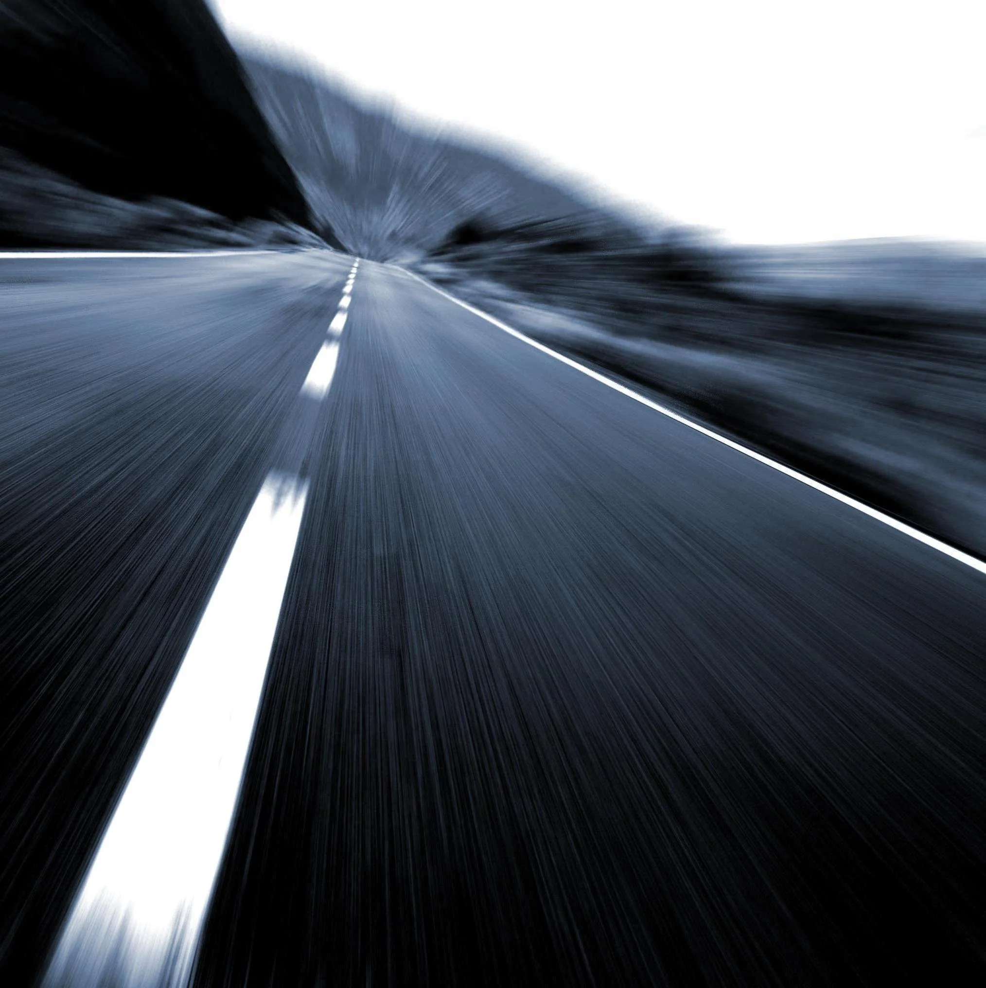 Blurred motion shot of a road with white dashed lines, surrounded by trees or landscape, taken from a moving vehicle.