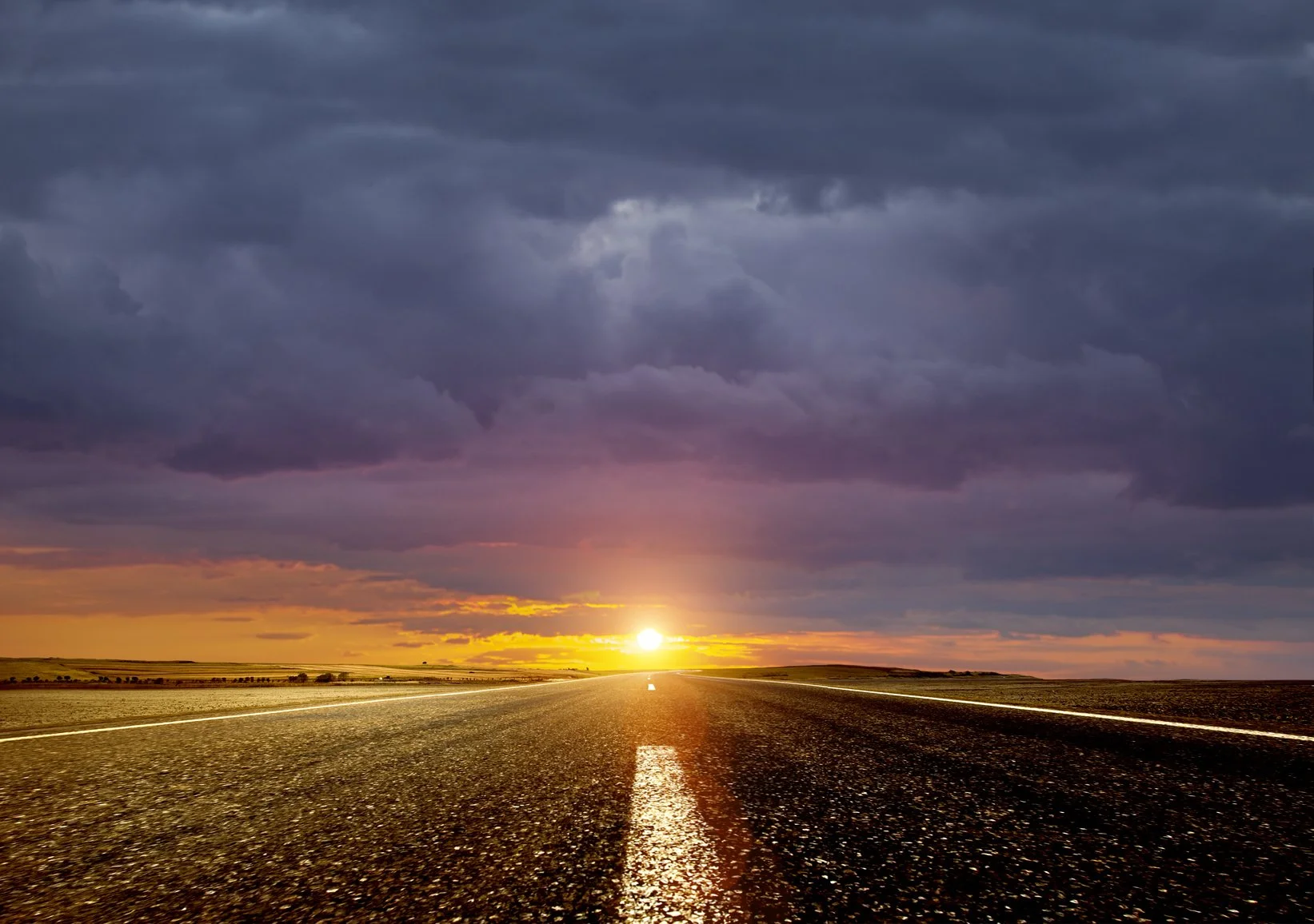 An empty road extending towards the horizon during sunset with dark clouds overhead.