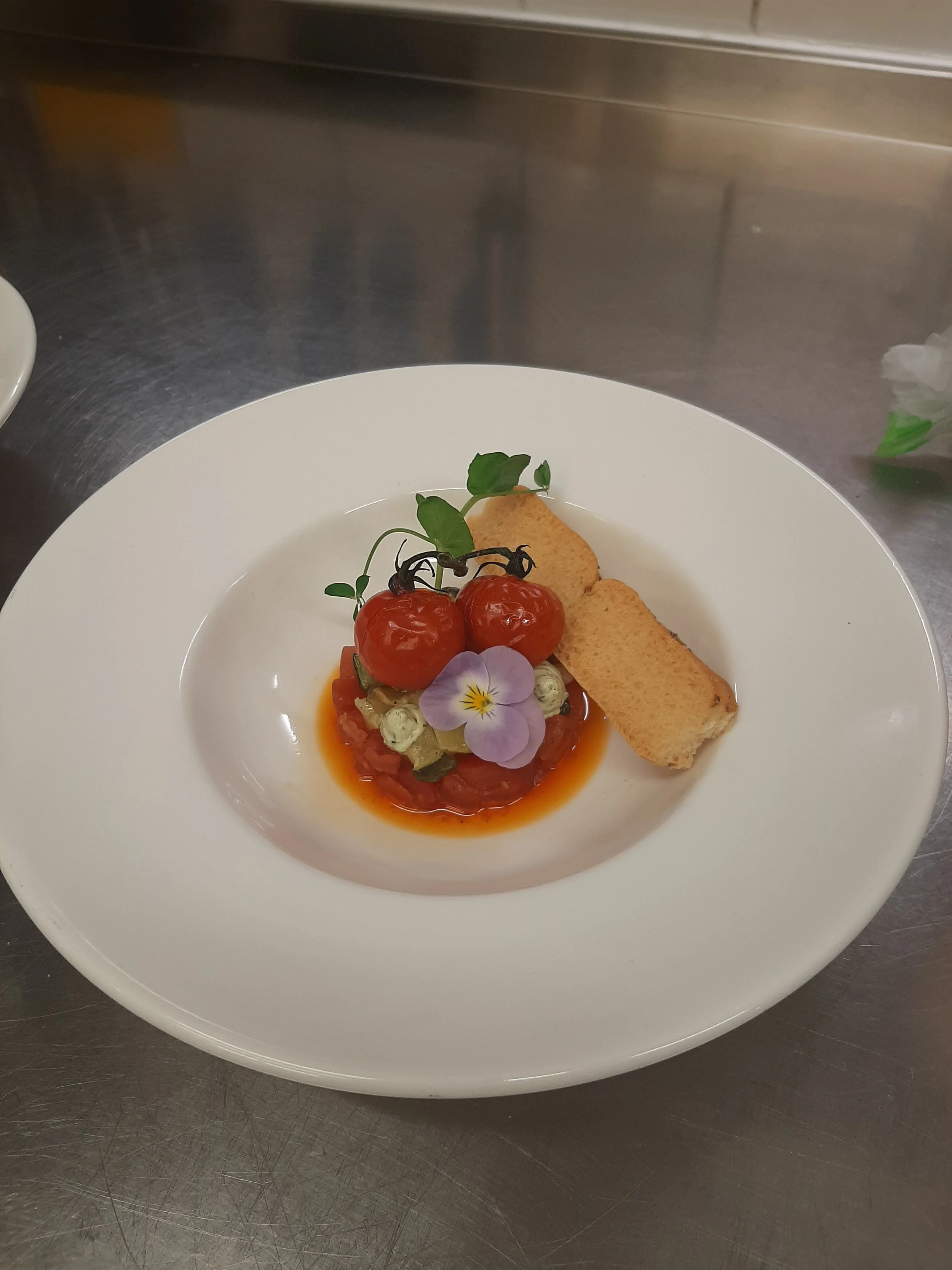 Gerecht met gepocheerde tomaat, een stuk toast, een bloemen- en kruidensamenstelling, geserveerd op een wit bord.