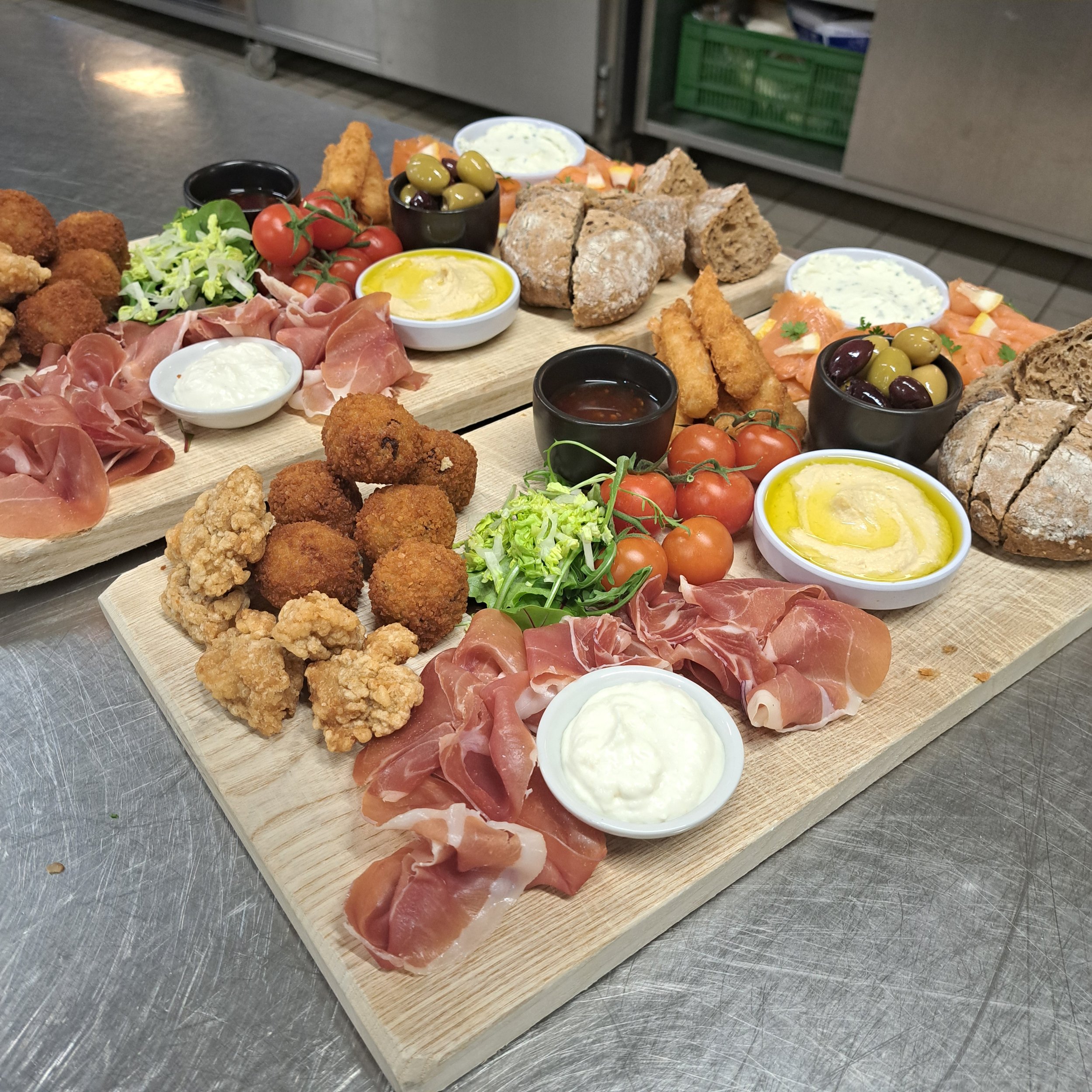 Elk type voedsel op houten planken, inclusief olijven, tomaten, prosciutto, kipnuggets, en diverse dipsauzen, met een metalen achtergrond.