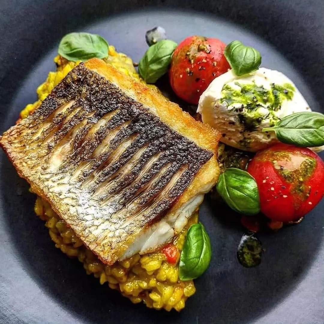Gebakken vis met ratatouille, geserveerd op een zwarte plaat met basilicumblaadjes en een bolletje kaas met pesto.