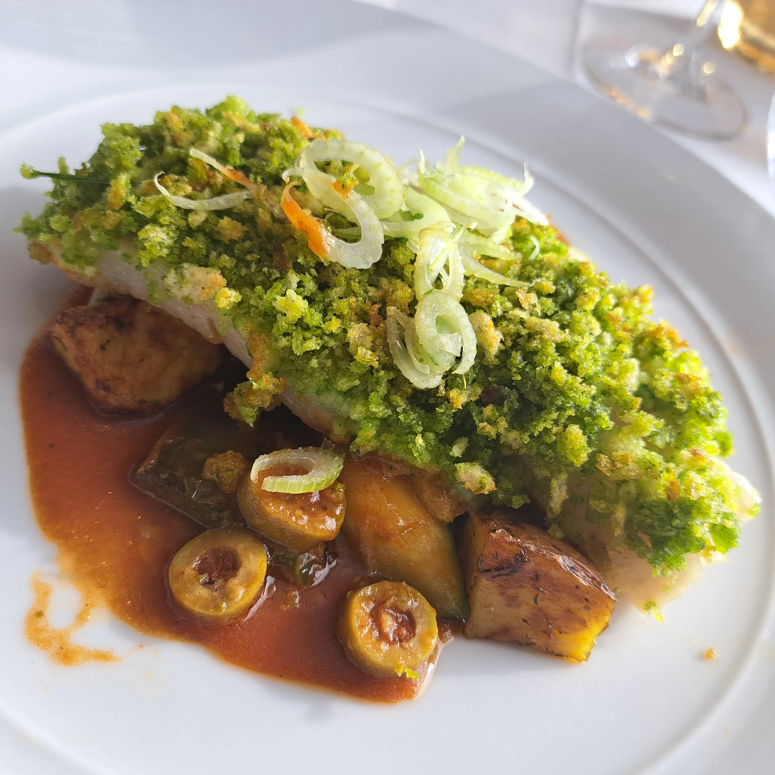 Een bord met gegrilde vis, bedekt met een groene kruidensaus, geserveerd met saus en aardappelen.