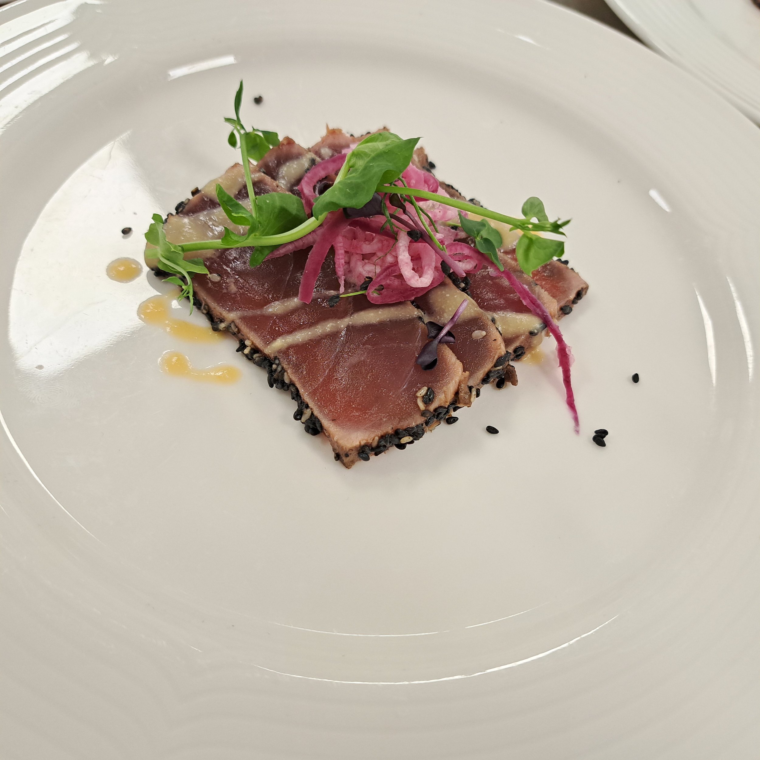 Gespikkelde ossenhaas carpaccio met rode ui, microgroenten, zwarte peper en een lichte olie op een wit bord.