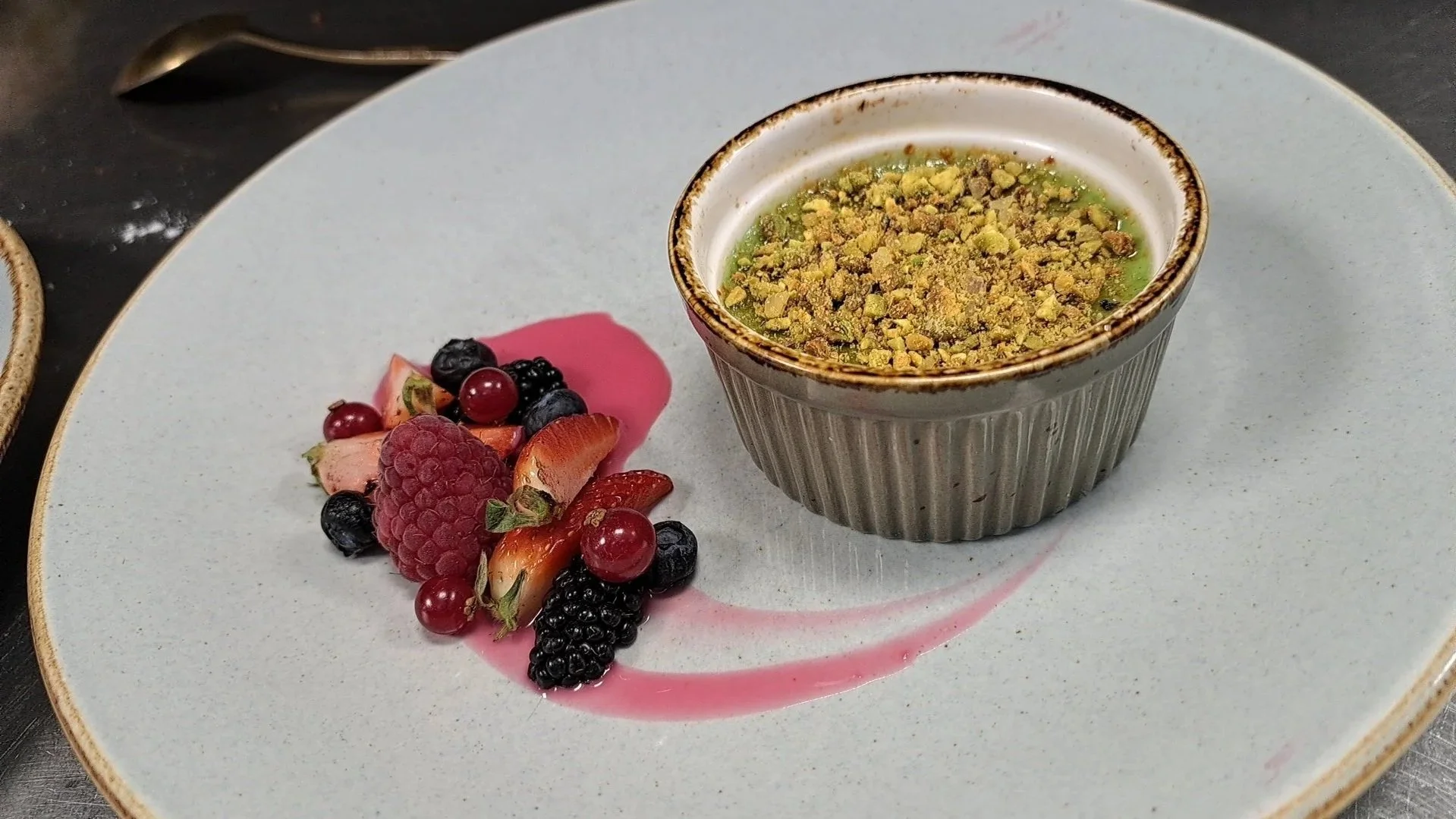 Een klein bakje met een groene crème of pudding bedekt met stukjes pistache nootjes, naast een beetje rood fruit en bessen op een wit bord met een roze saus en een lepel op de achtergrond.