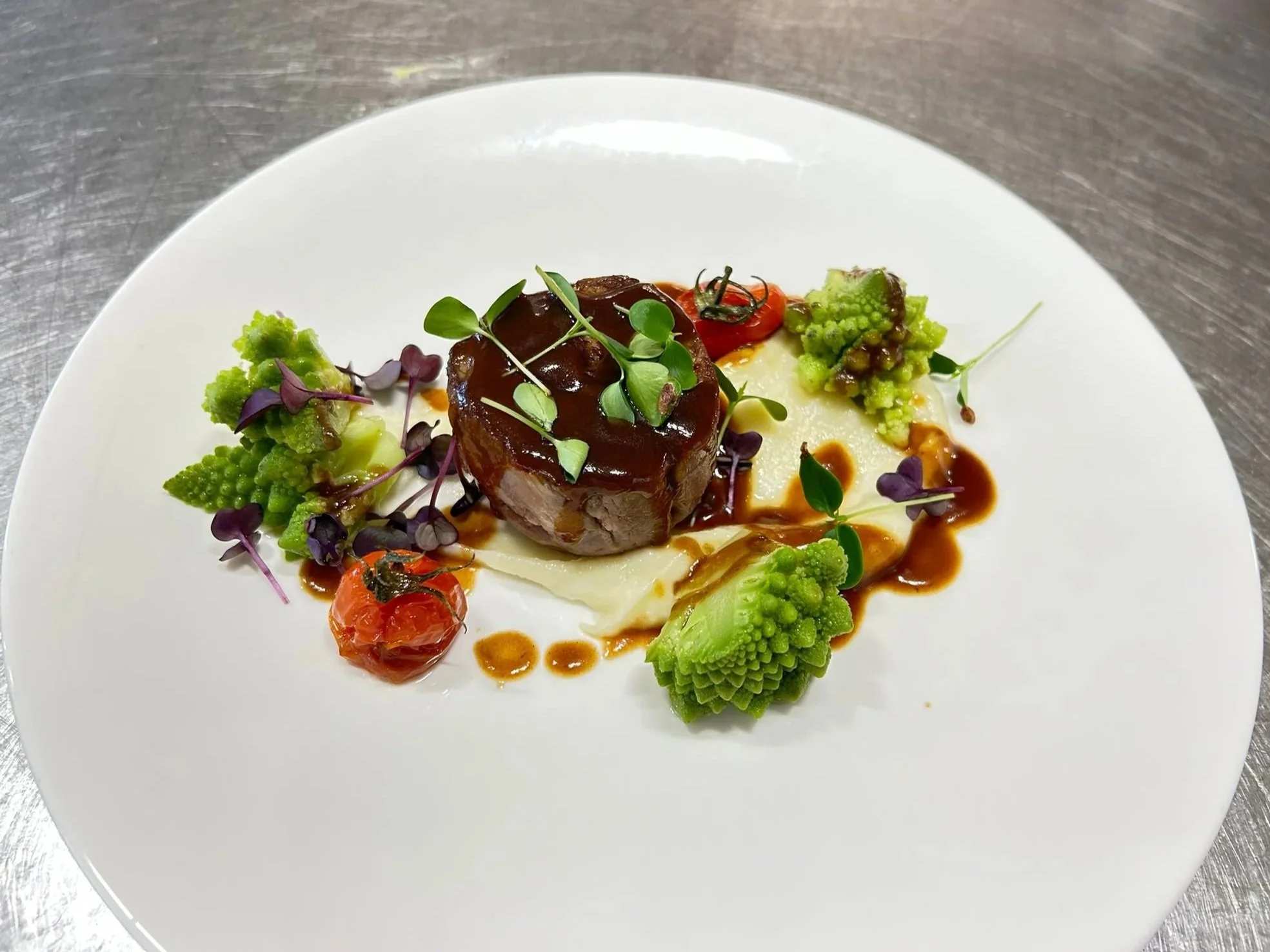 Gerecht met vlees, groenten, microgroenten en saus op een wit bord, gelegen op een metalen tafel.