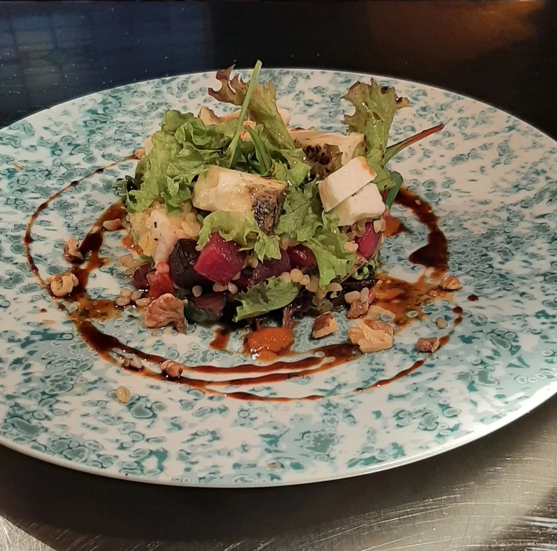 Gerecht met salade, gegrilde groenten, en een saus op een blauw-wit mokkakleurige bord.